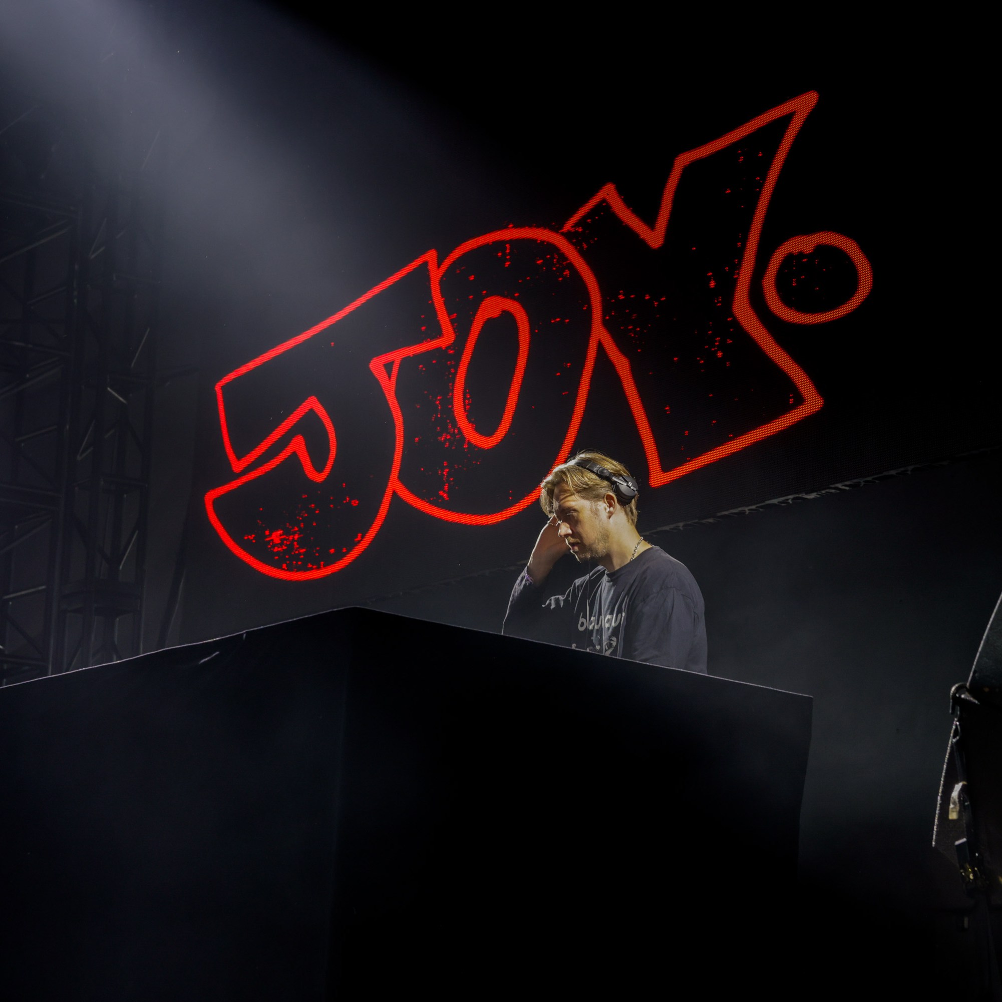 A DJ stands focused at his turntable, illuminated under stage lights, with the word &quot;Joy&quot; in large, bright red letters behind him.