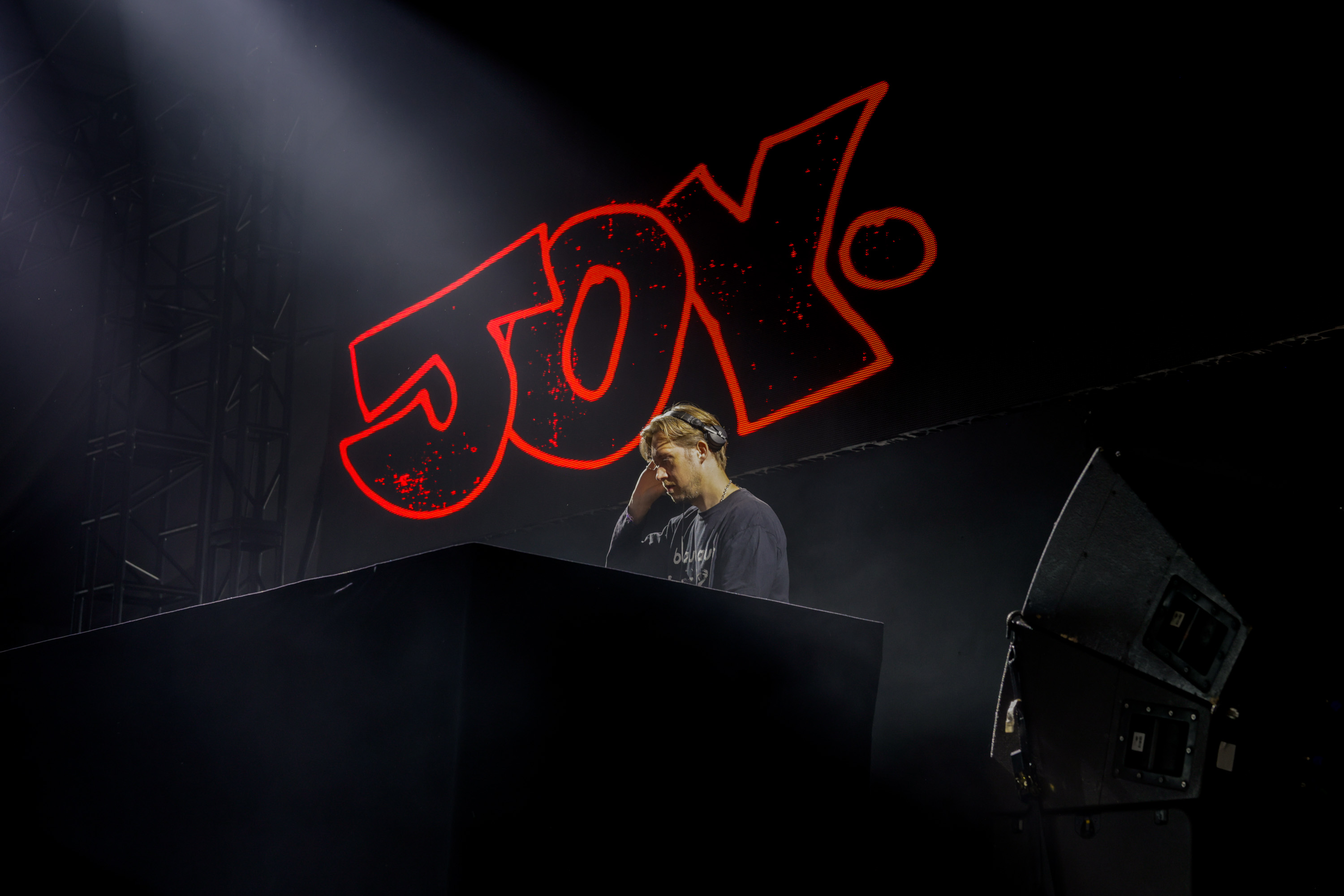 A DJ stands focused at his turntable, illuminated under stage lights, with the word &quot;Joy&quot; in large, bright red letters behind him.