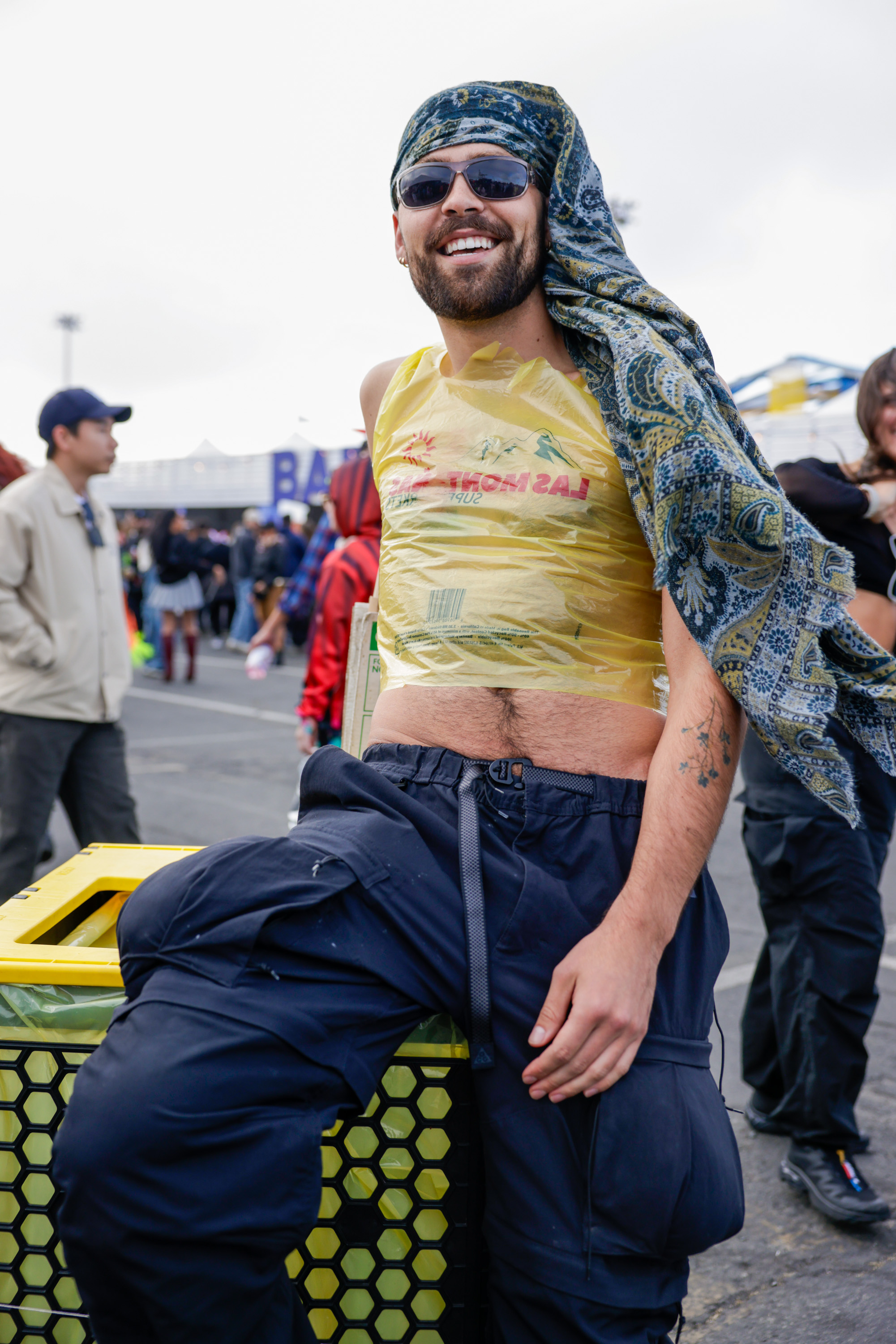 A bearded man in sunglasses wears a headscarf and a yellow plastic bag as a top, leaning on a trash bin at an outdoor event, smiling brightly.