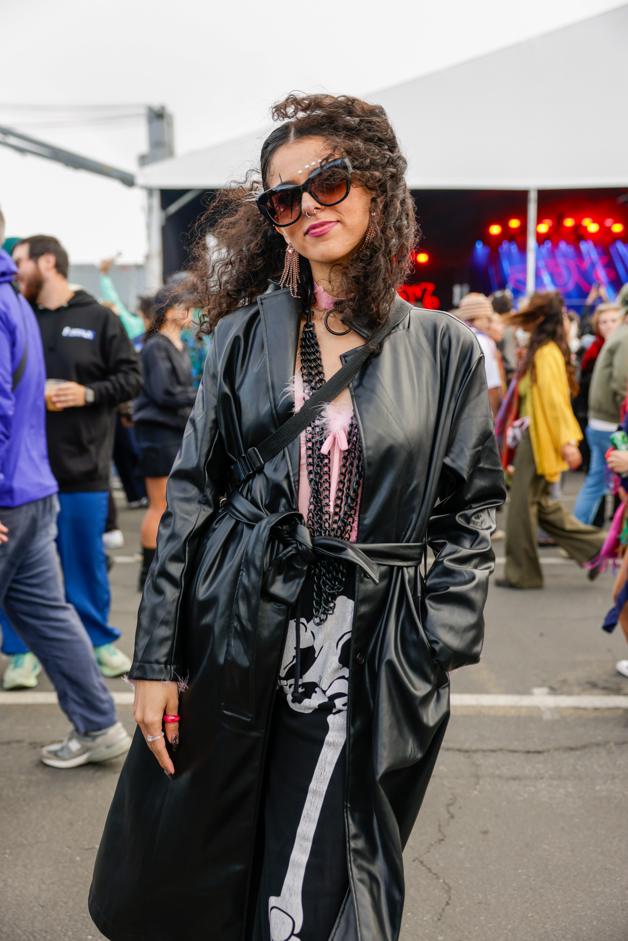 A woman with curly hair wearing a black leather coat, large sunglasses, and earrings stands at an outdoor event with crowds and a stage in the background.