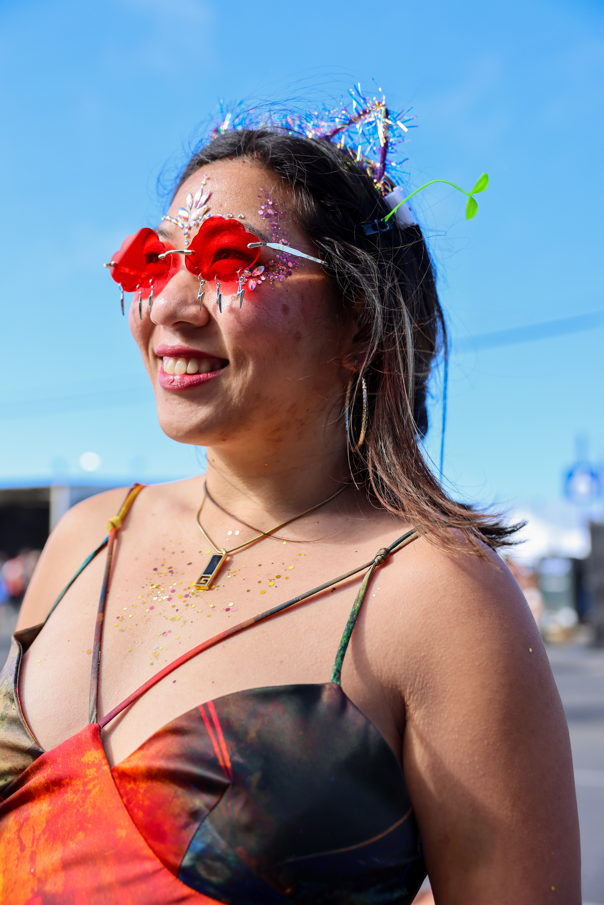 A woman wearing colorful, heart-shaped glasses and a sparkly tiara smiles under a blue sky. She is adorned with glitter on her face and chest, and wears a vibrant dress.