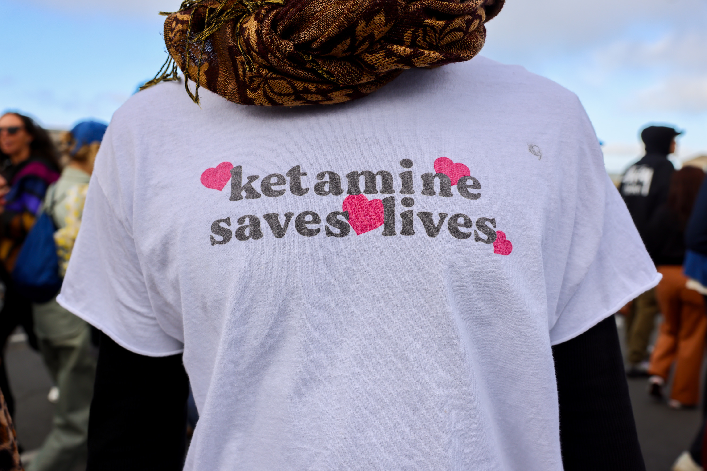 The image shows a person wearing a white T-shirt that says &quot;ketamine saves lives&quot; with pink heart shapes around the text. Other people are blurred in the background.