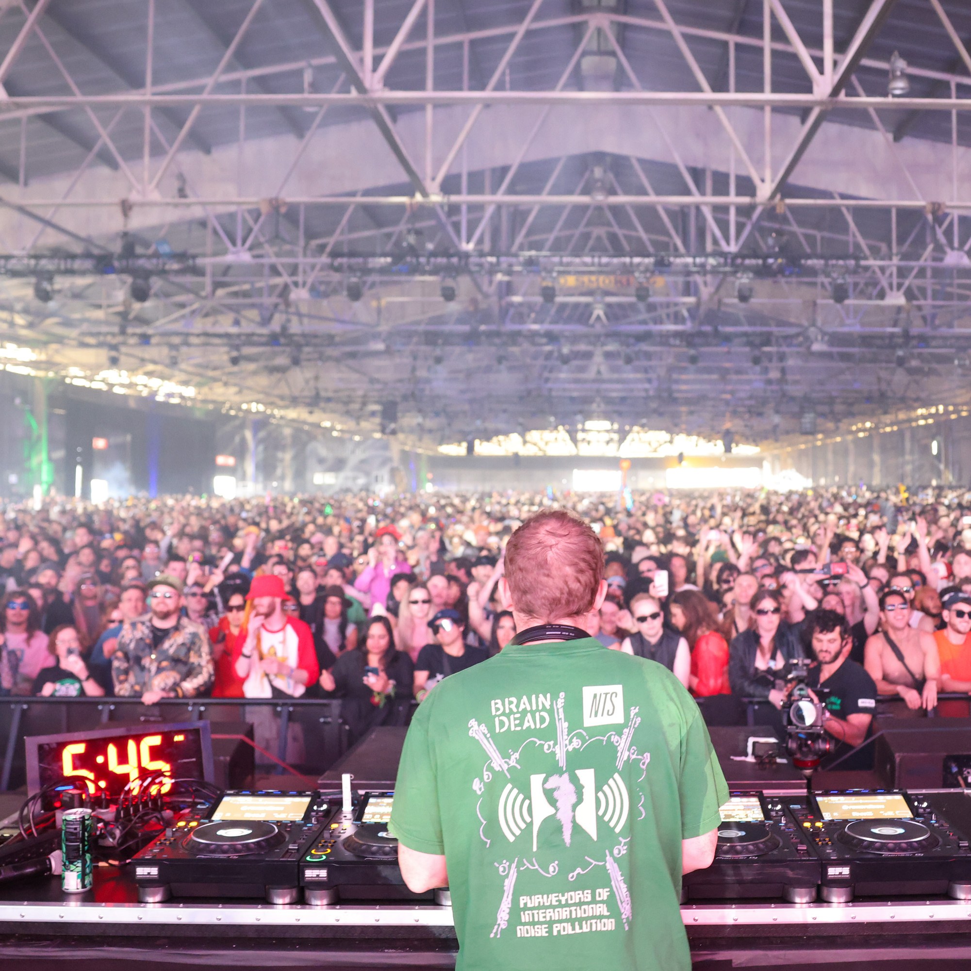 A DJ stands at a console in front of a massive crowd in an indoor venue. The audience is packed, energetic, and many people are wearing sunglasses and colorful attire.