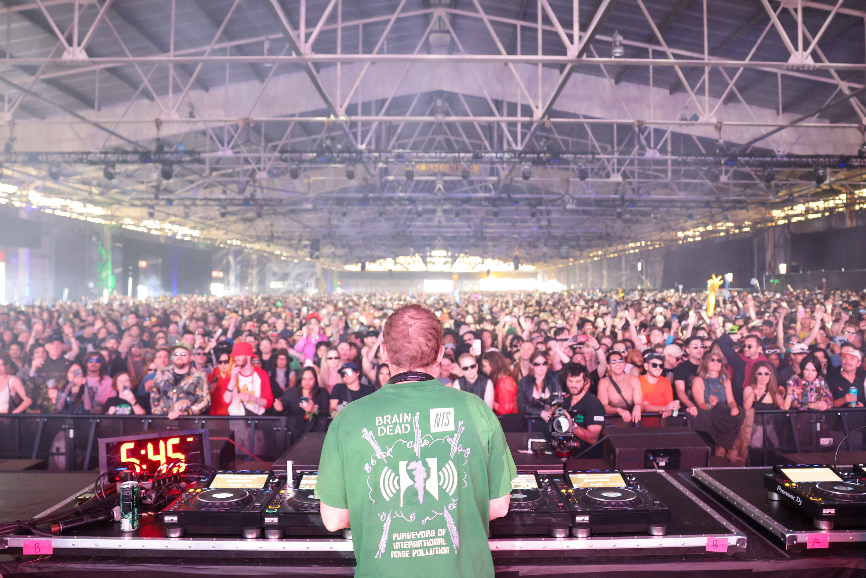 A DJ stands at a console in front of a massive crowd in an indoor venue. The audience is packed, energetic, and many people are wearing sunglasses and colorful attire.