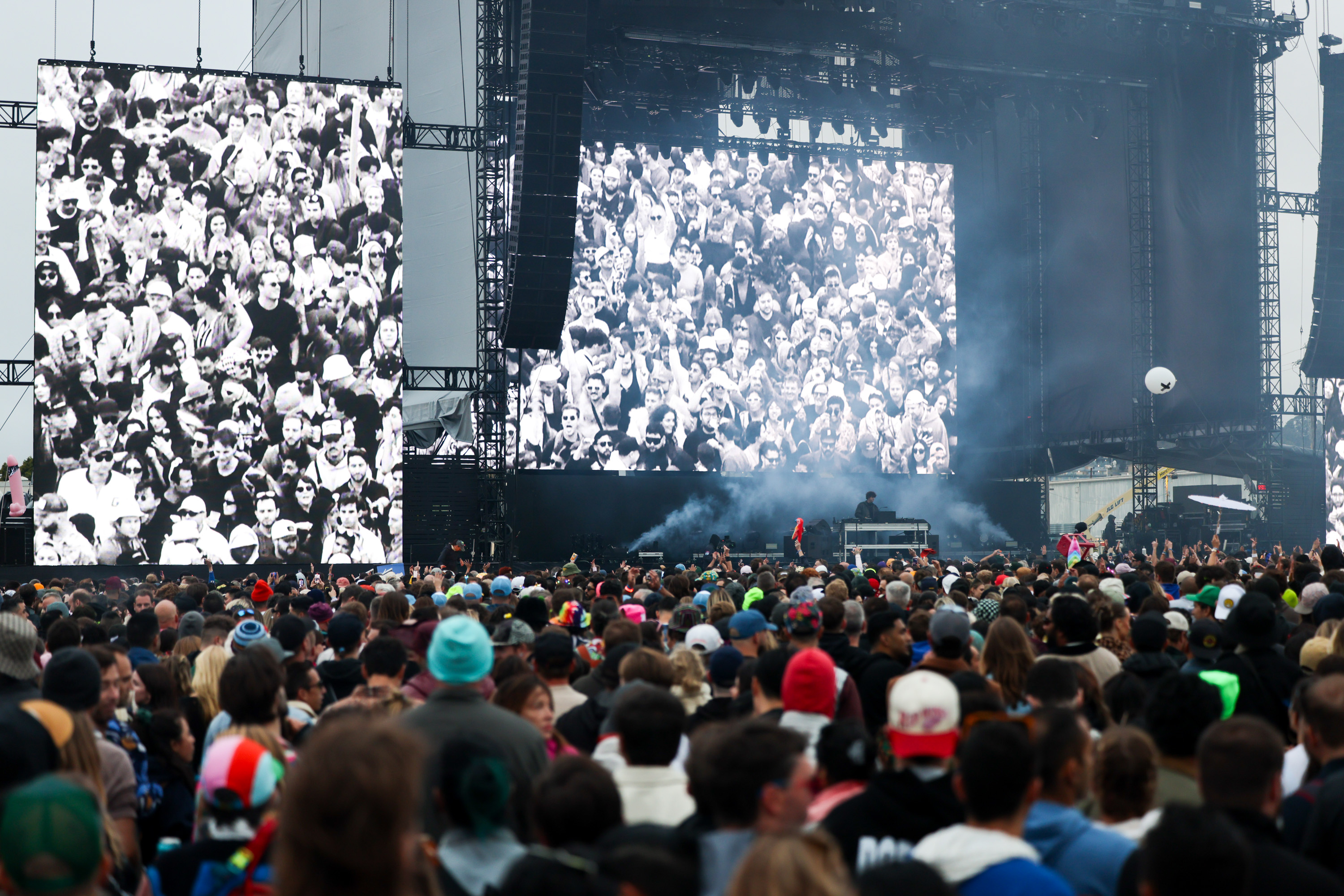 A large crowd faces a concert stage with a DJ. Two big screens show black-and-white crowd visuals, and there's a visible haze of smoke.