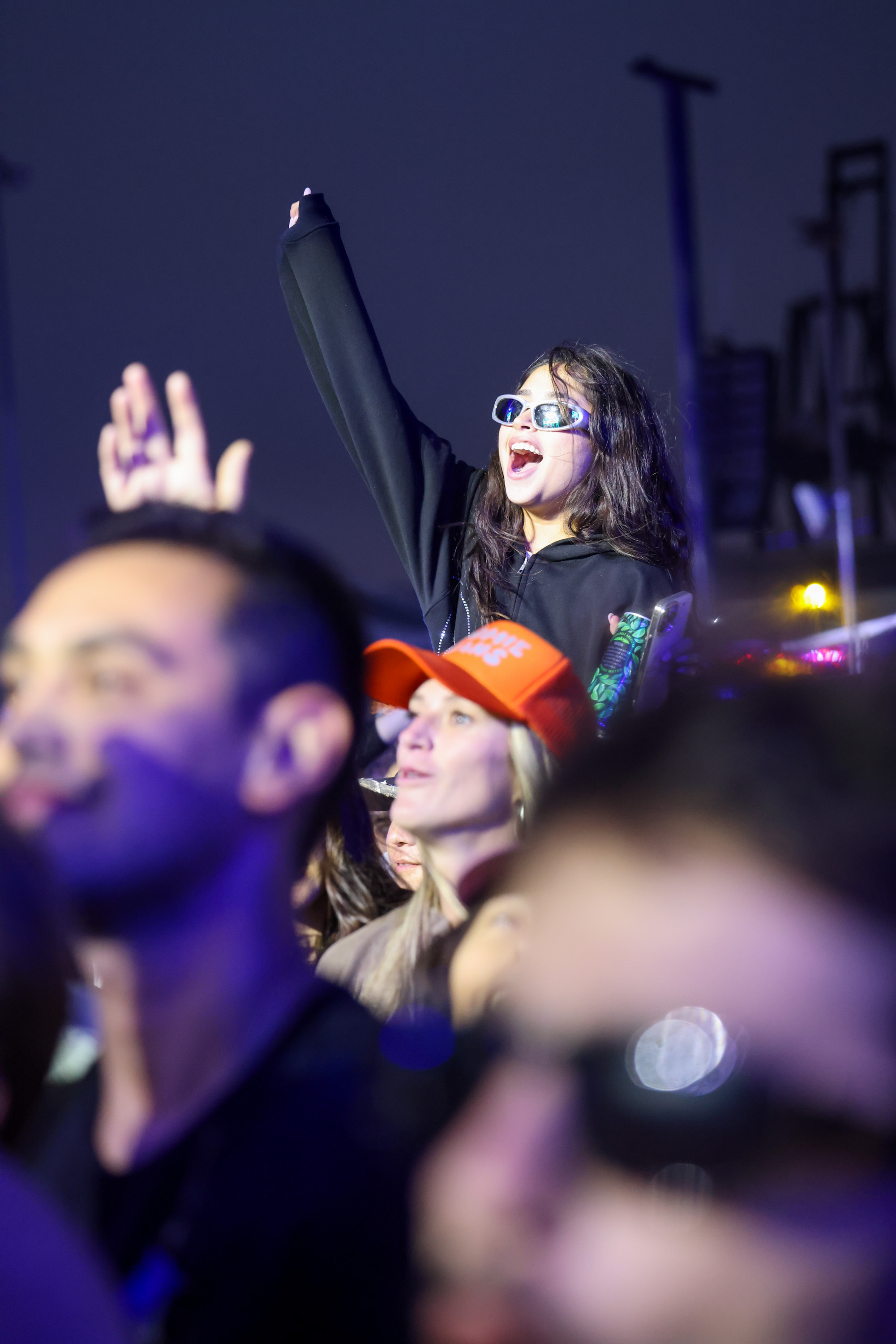 A crowd of people at a nighttime event, with a young woman wearing sunglasses and a black hoodie energetically raising her hand while others look on.