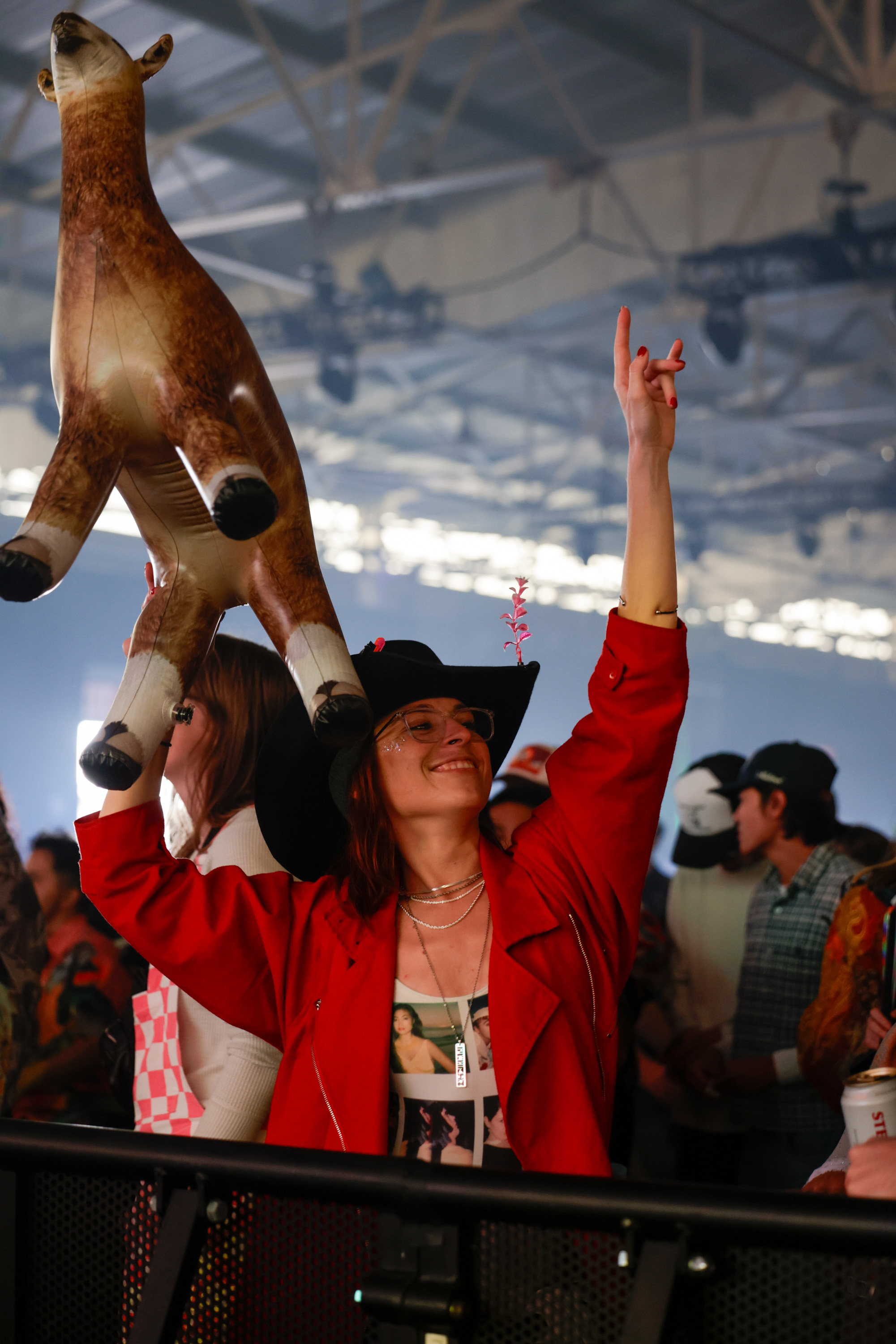 A joyful person in a red jacket and black hat holds an inflatable llama above their head, surrounded by a crowd in a large, high-ceilinged venue.