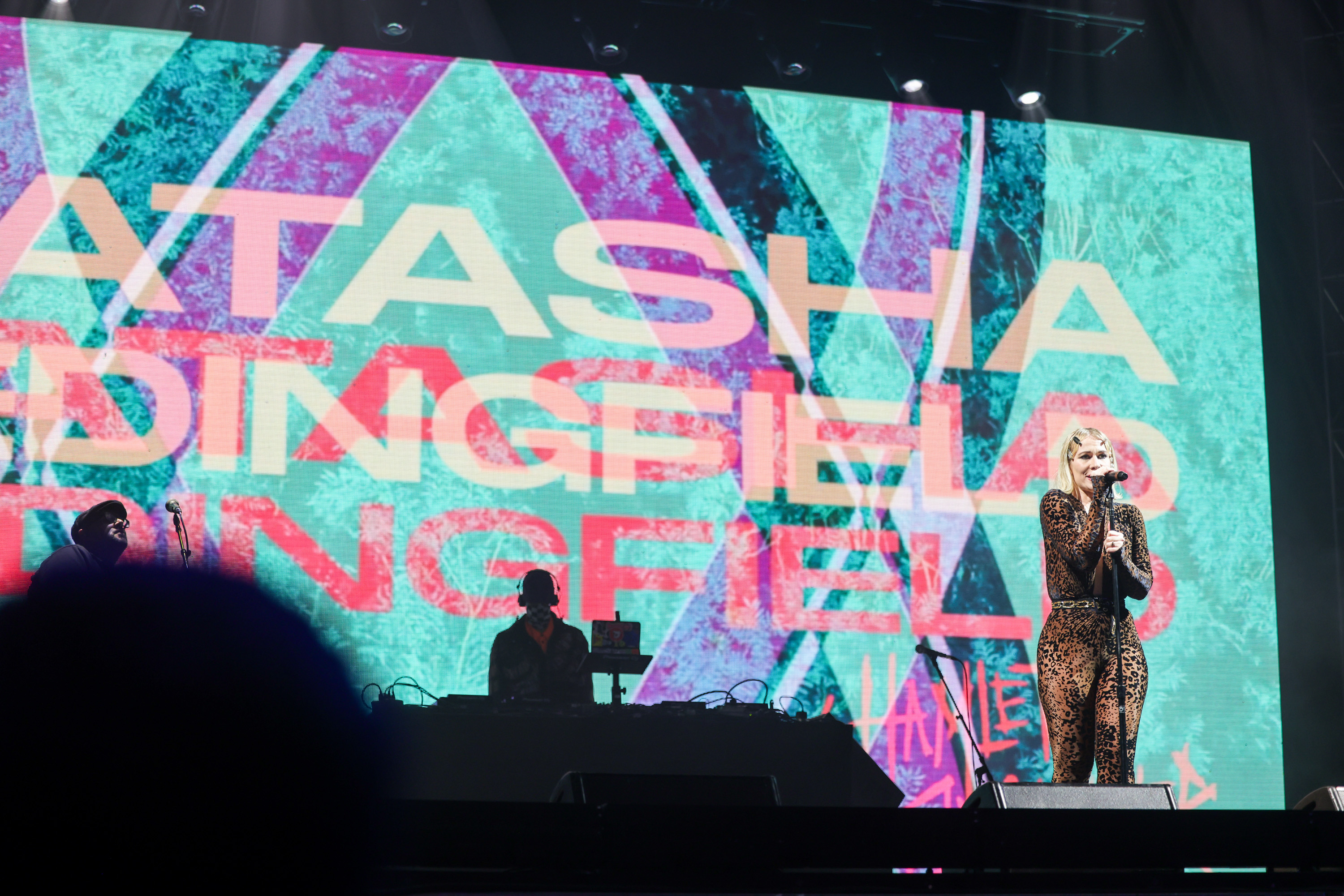 A performer in a leopard-print outfit sings into a microphone on stage. A large screen behind displays colorful graphics and the text &quot;NATASHA BEDINGFIELD.&quot;