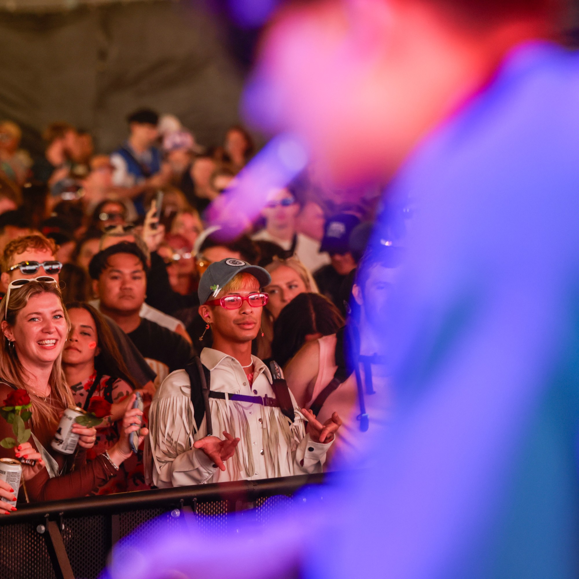 A colorful crowd at a concert leans against a barrier, enjoying the show and smiling, many wearing sunglasses and holding drinks, with the performer blurred in the foreground.