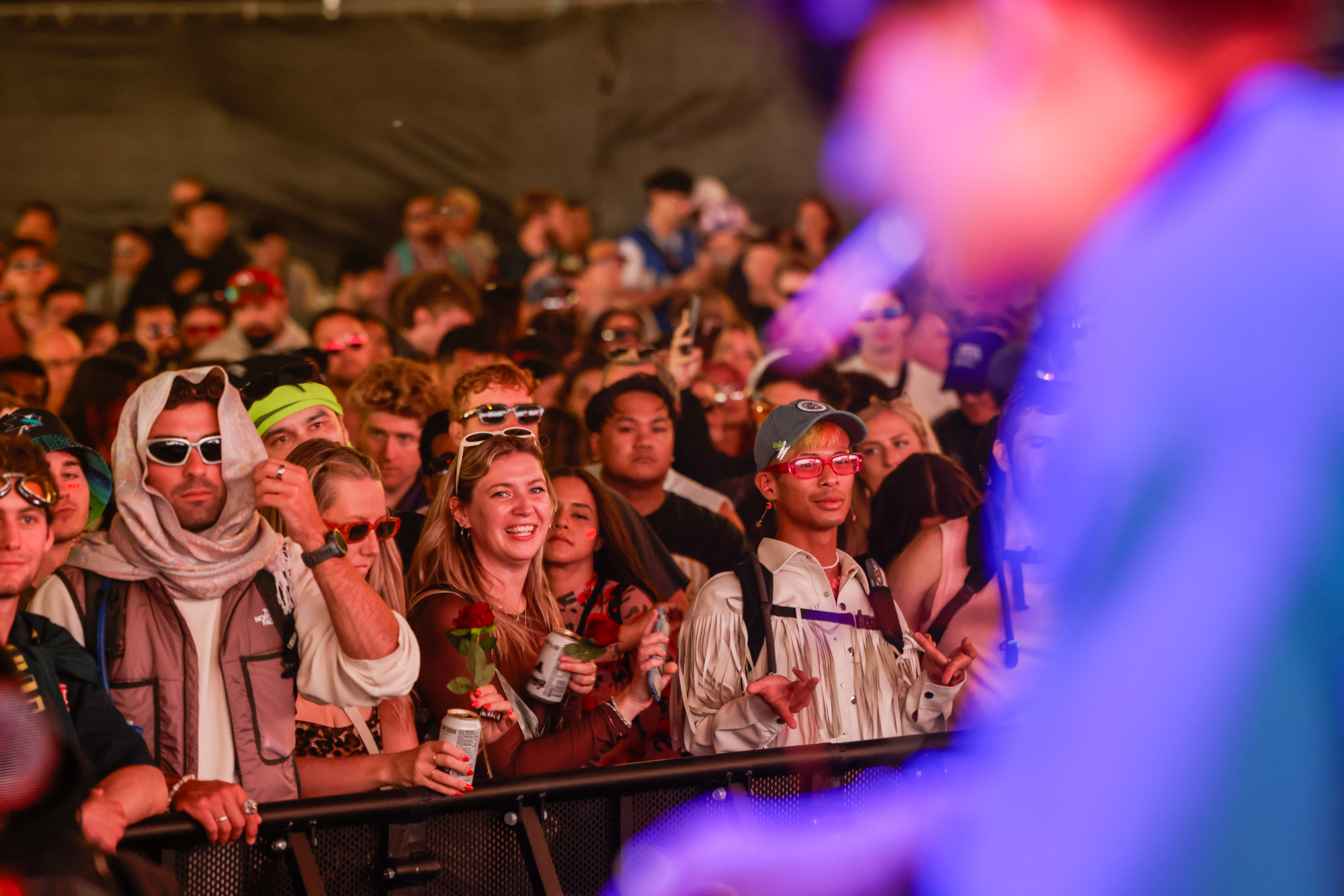 A colorful crowd at a concert leans against a barrier, enjoying the show and smiling, many wearing sunglasses and holding drinks, with the performer blurred in the foreground.