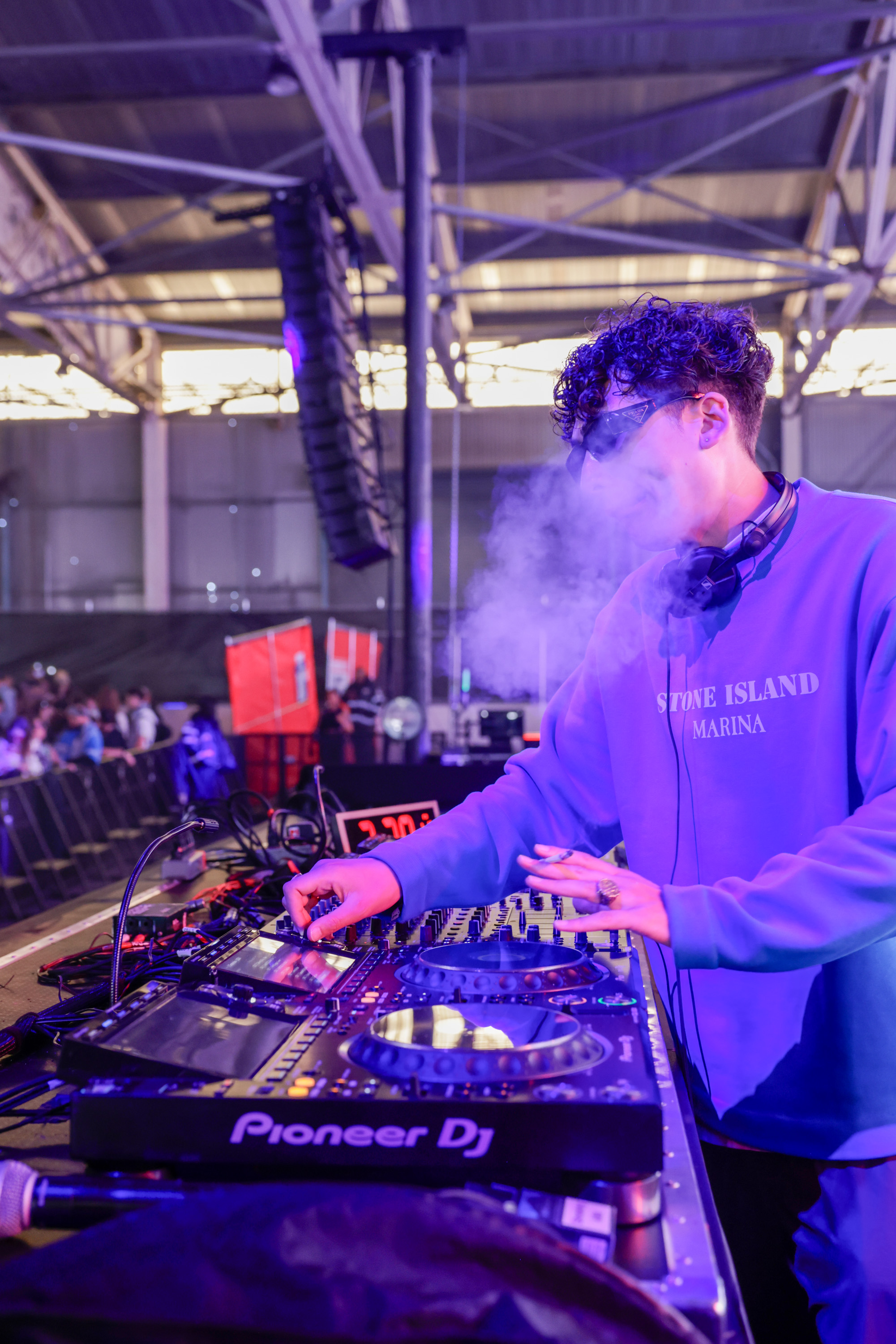 A DJ in a purple sweater, surrounded by fog, performs on a stage with a Pioneer DJ setup. He wears sunglasses and headphones, and an audience is visible in the background.