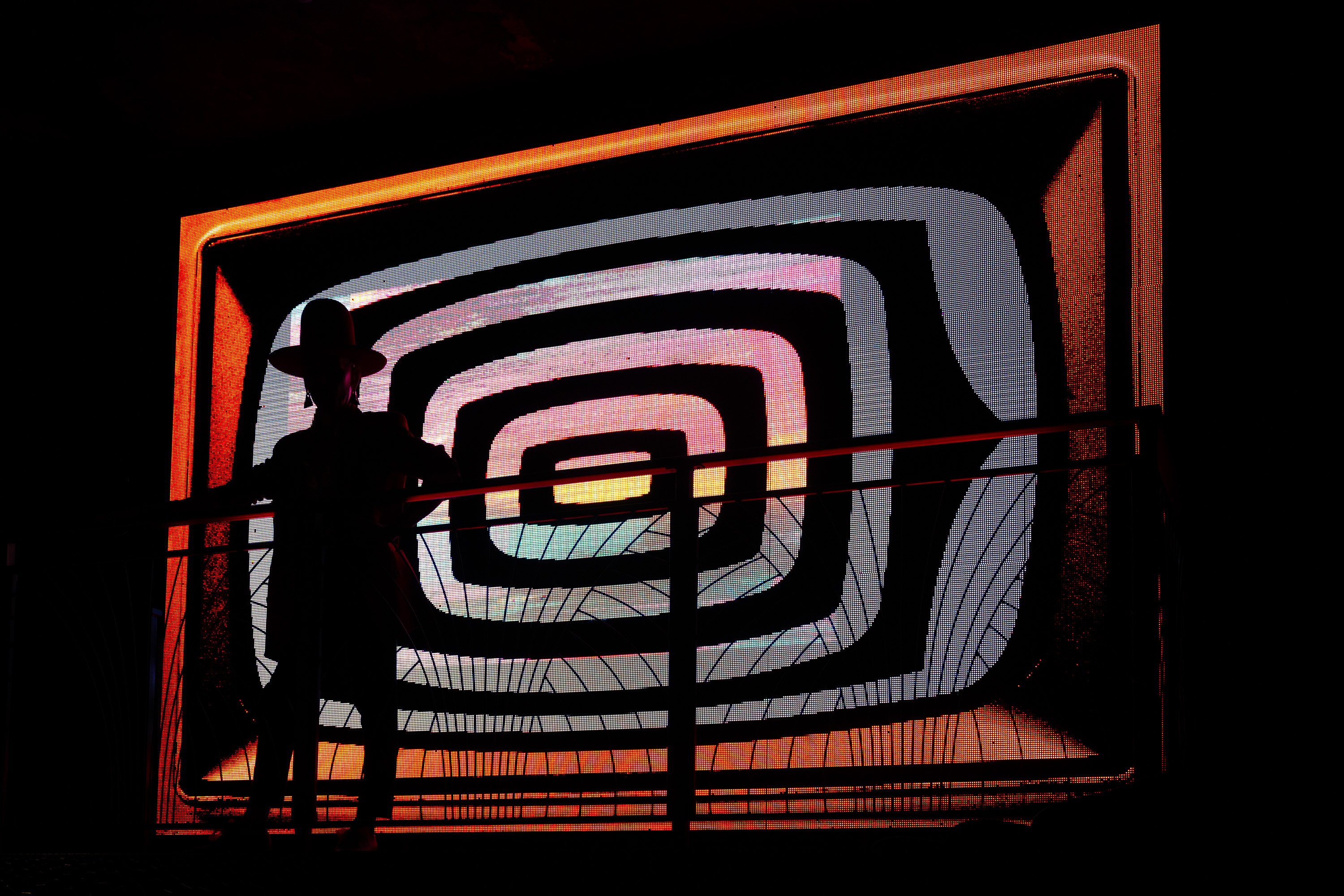 A silhouetted person wearing a hat stands in front of a large, hypnotic, spiral-patterned, colorful screen, creating an artistic and dramatic visual.