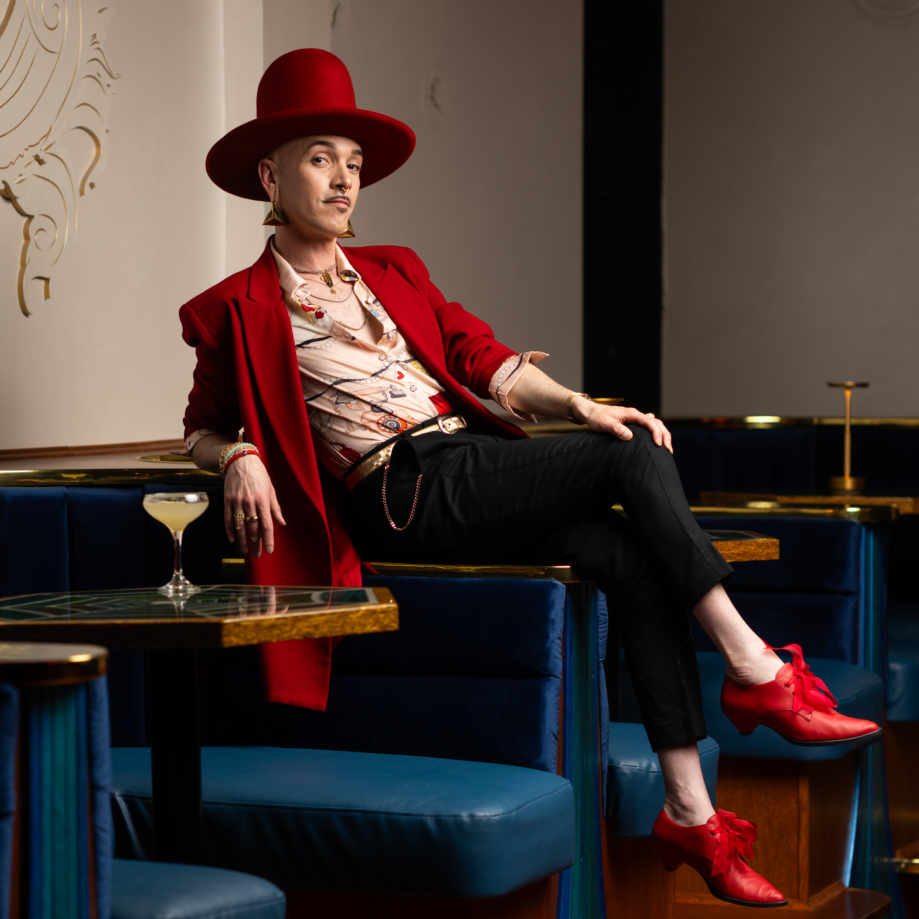 A person in a large red hat, red jacket, and red shoes lounges on a blue upholstered bench. They wear a patterned shirt and black pants, with a cocktail nearby.