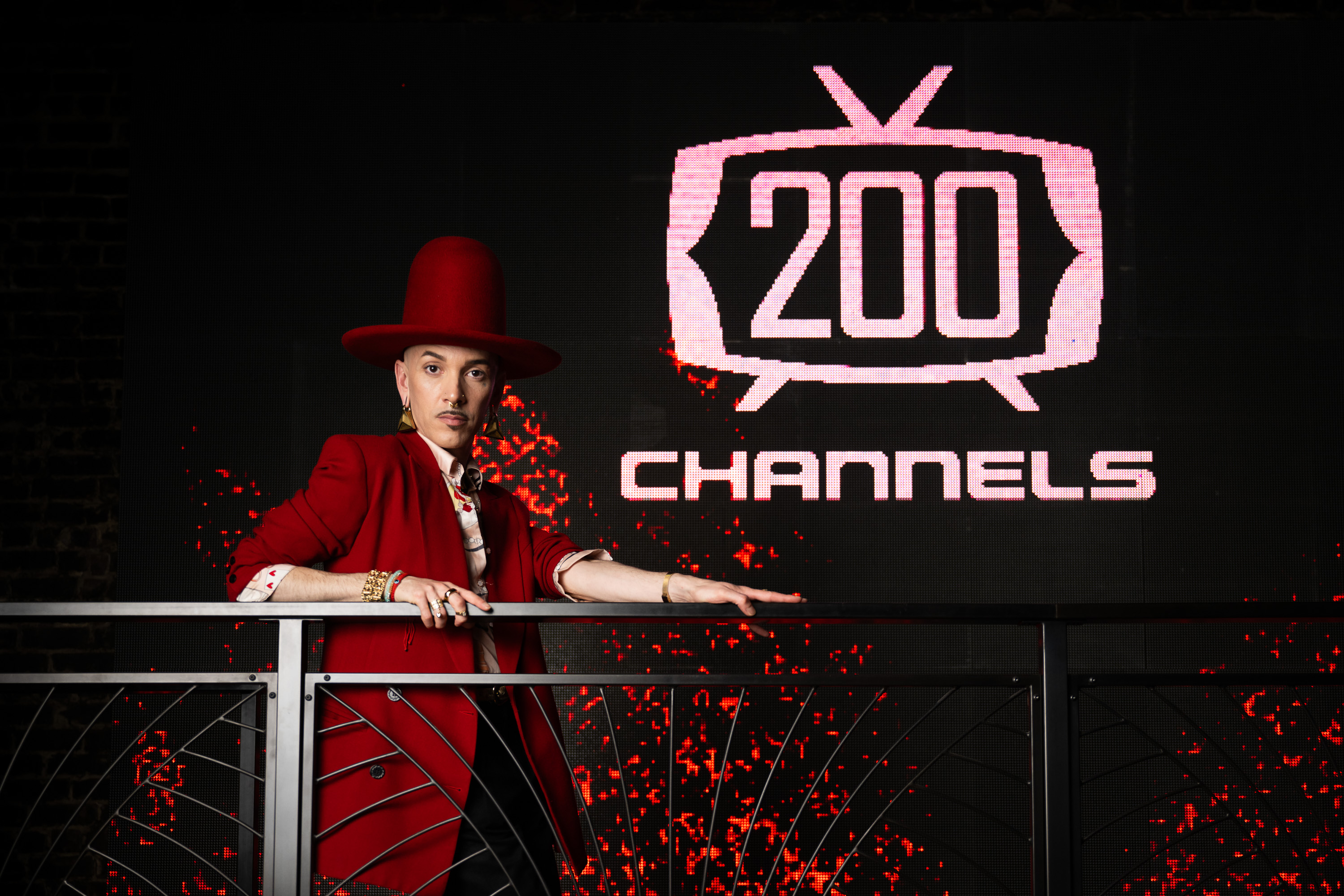 A person in a red outfit and large red hat stands against a backdrop with &quot;200 Channels&quot; displayed on a screen, leaning on a modern railing.