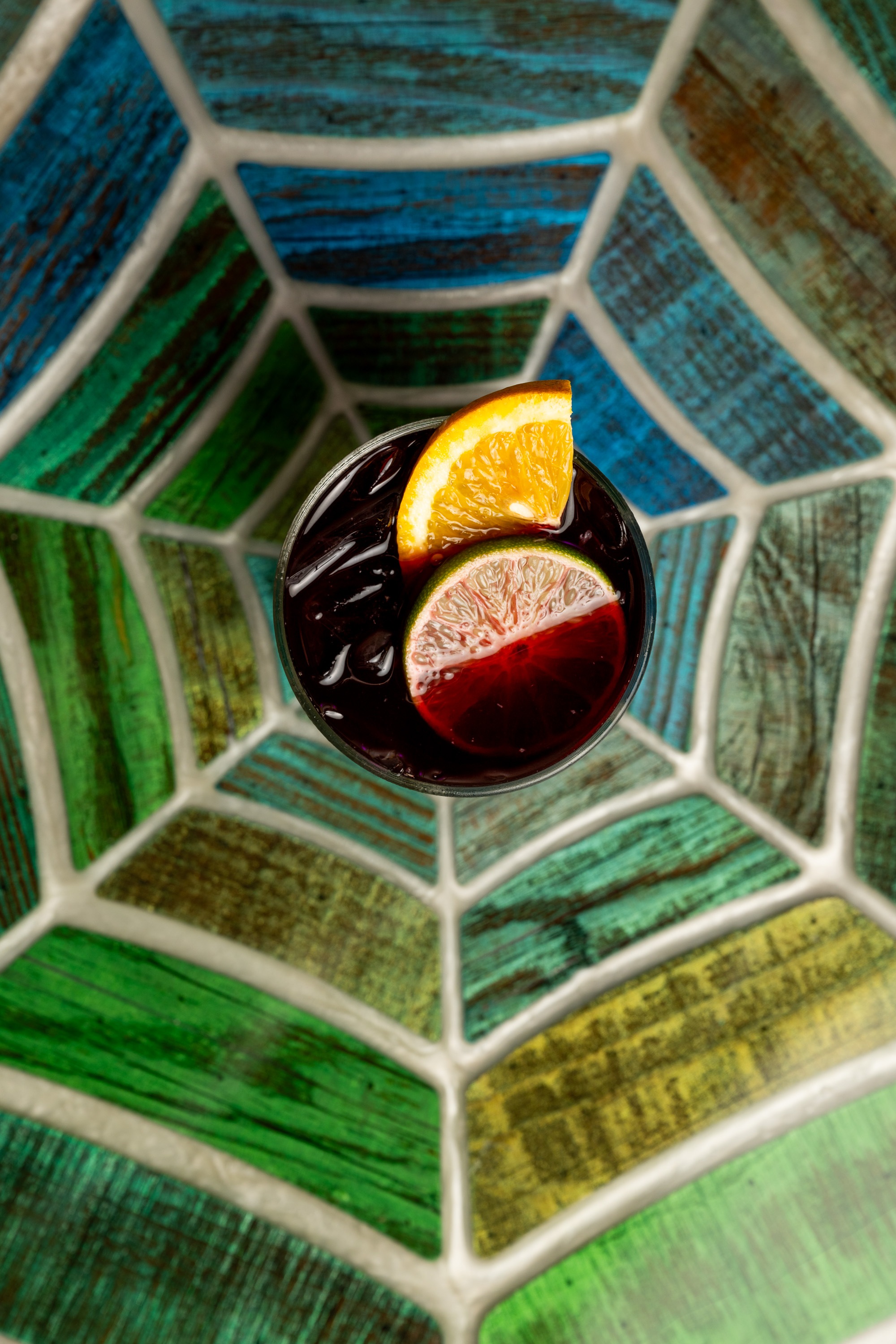 A dark red drink in a clear glass is garnished with orange and lime slices, set against a vibrant, mosaic-patterned background with shades of green and blue.