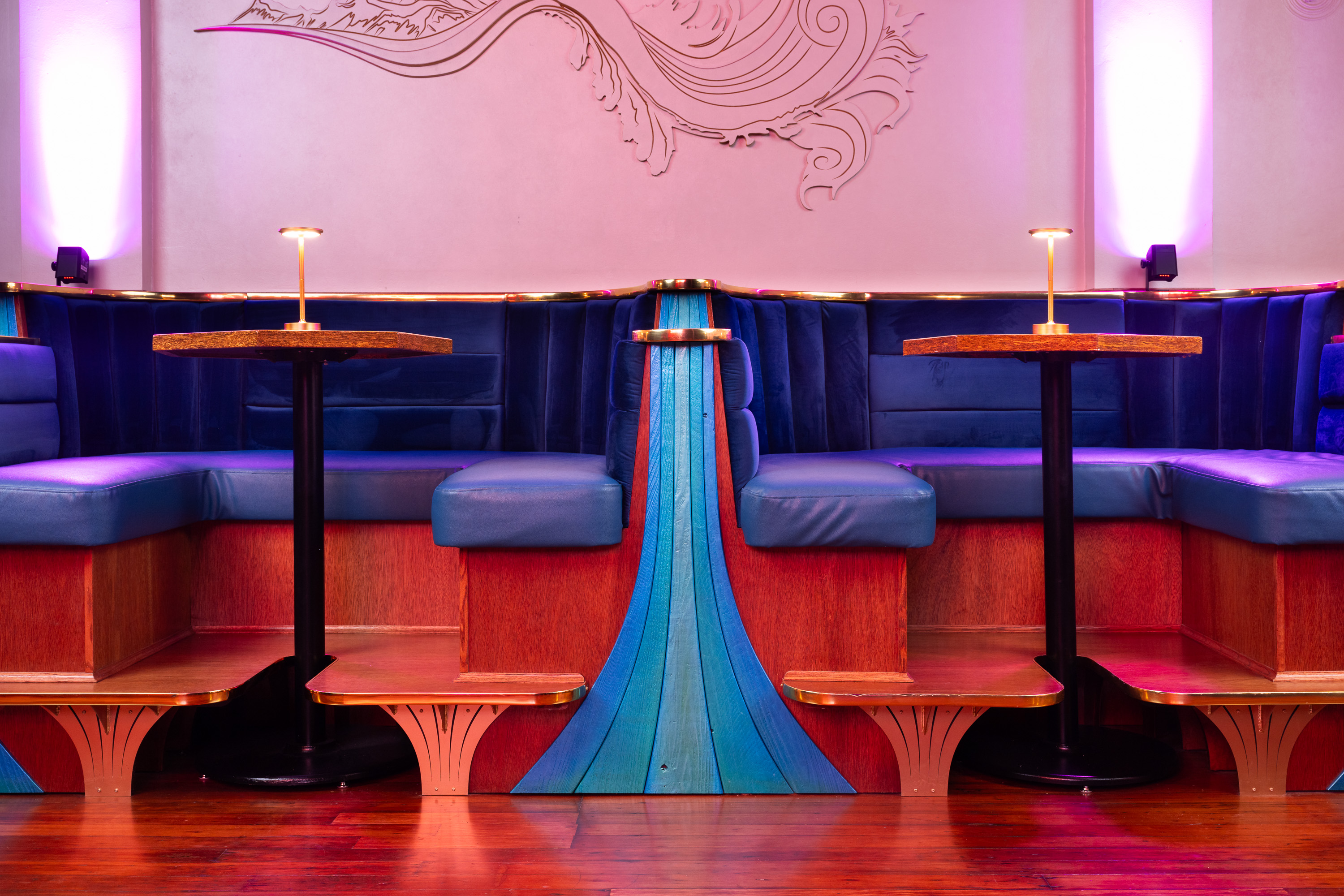 The image shows a plush, blue velvet booth seating area with wooden tables and yellow lamps, under pink ambient lighting, and intricate wall decor above.