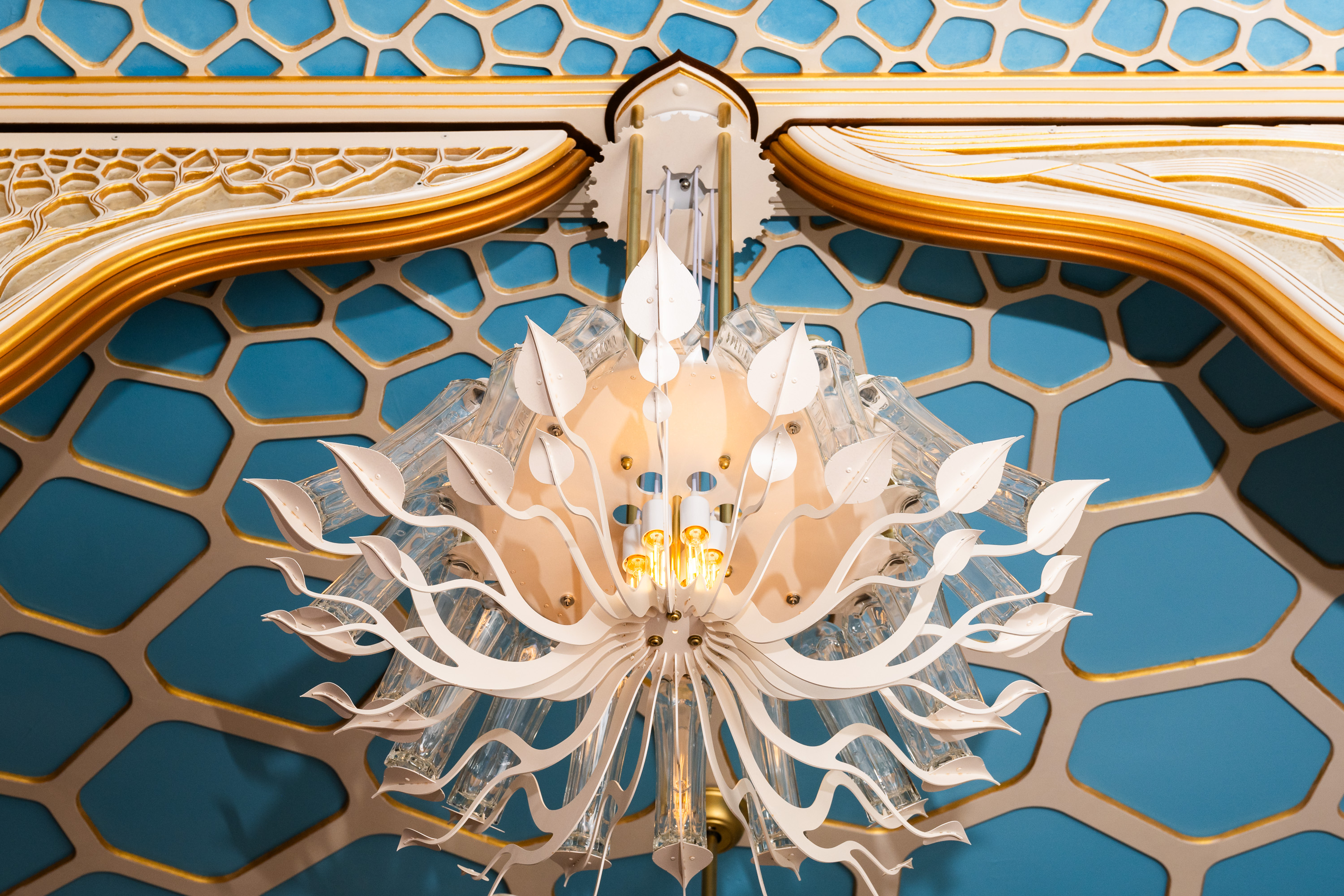 The image features an ornate, modern chandelier with white, leaf-like decorations against a blue, hexagonal-patterned ceiling with gold accents.