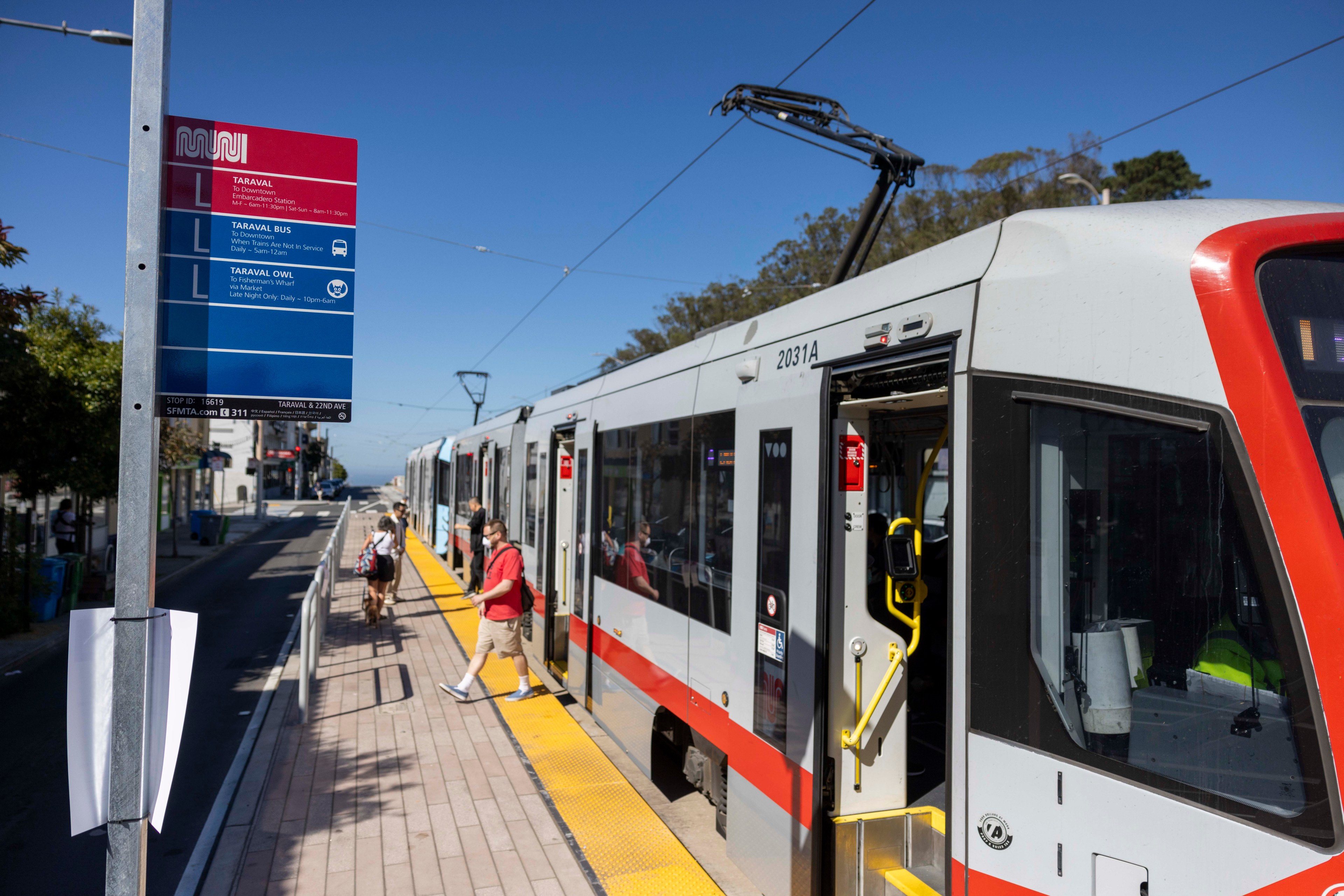 A white and red train is stopped at a station, with its doors open. Several passengers are boarding. A nearby pole displays a "Muni" sign with route information.