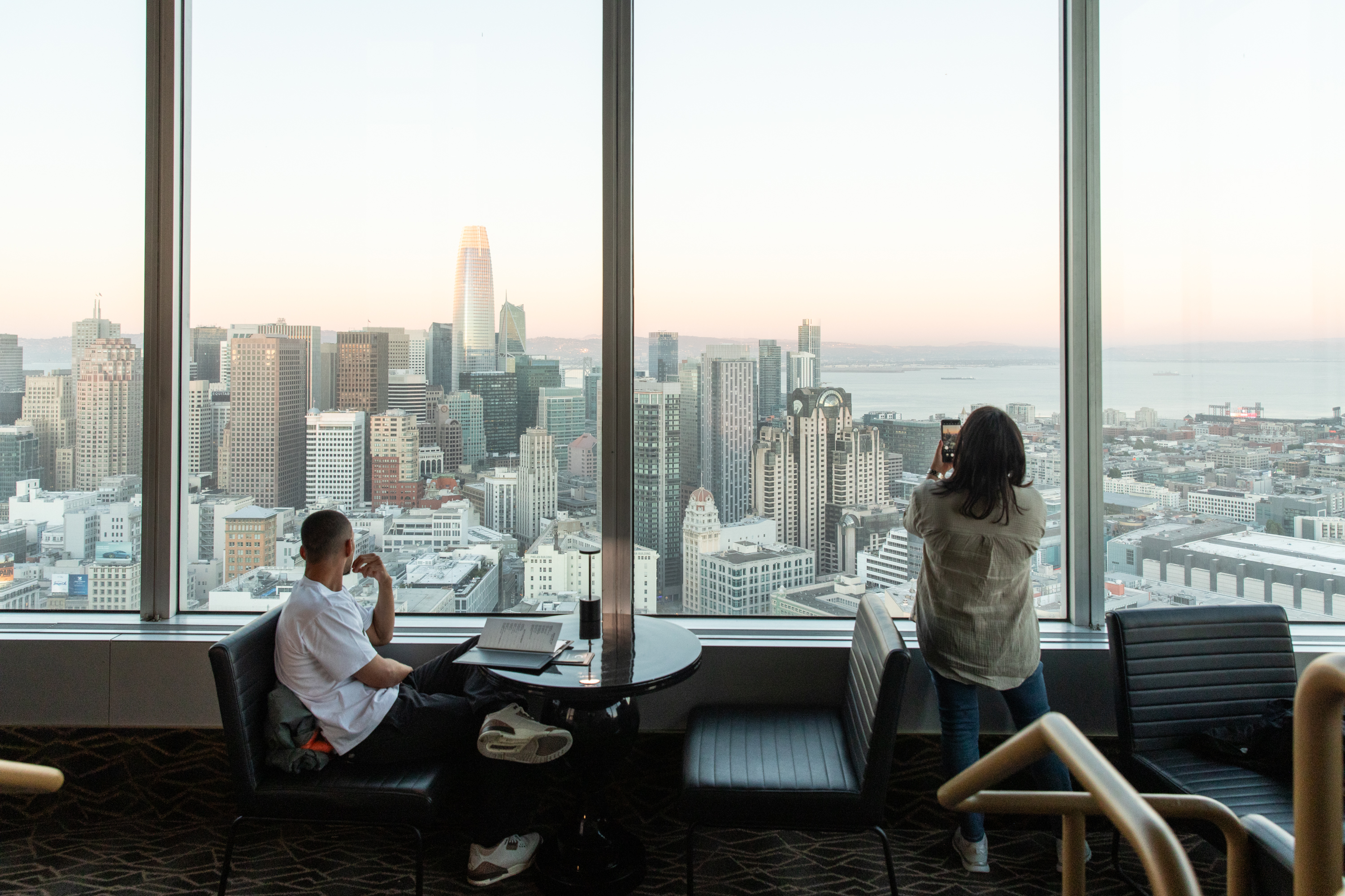 A man and a woman are in a lounge area with large windows overlooking a city skyline. The man is seated, looking out, while the woman stands, taking a photo.