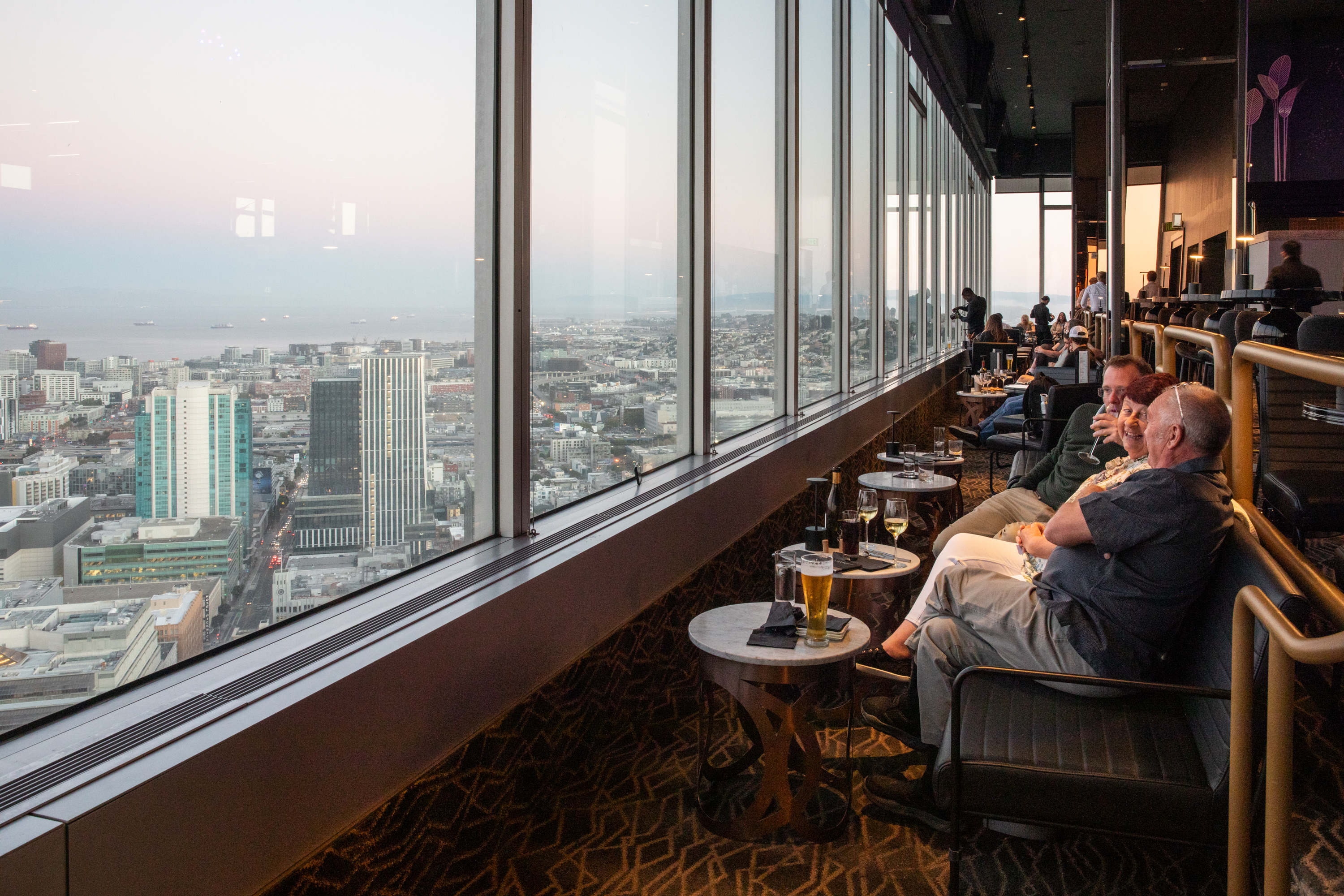 People are relaxing at a high-rise bar or restaurant with large windows, enjoying drinks and a panoramic city and ocean view at sunset.