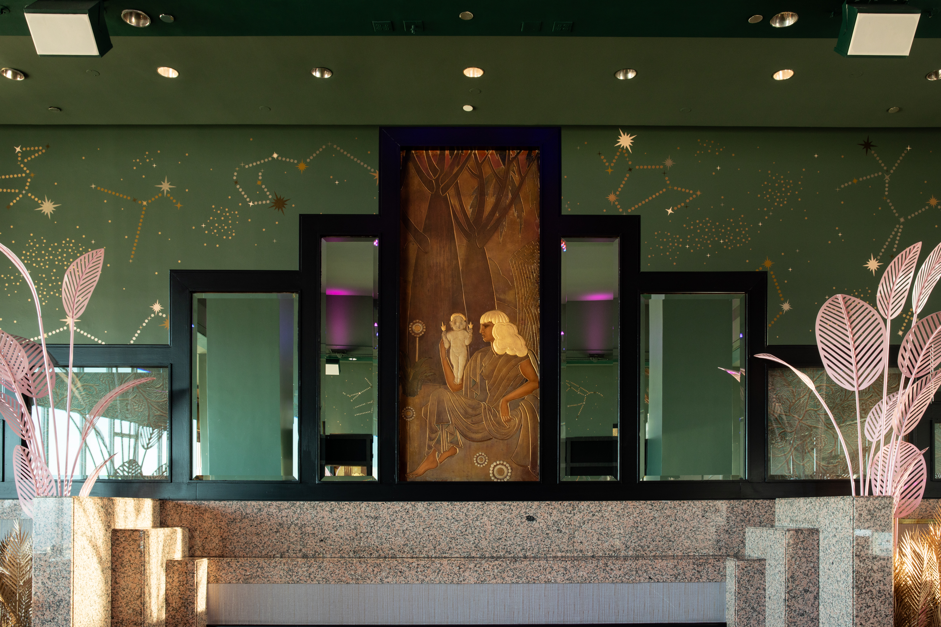 The image shows a decorative wall with a bronze-colored mural of a woman and child in the center, framed by geometric mirrors. The backdrop features green walls adorned with golden constellations, and pink metal plant sculptures flank the sides.