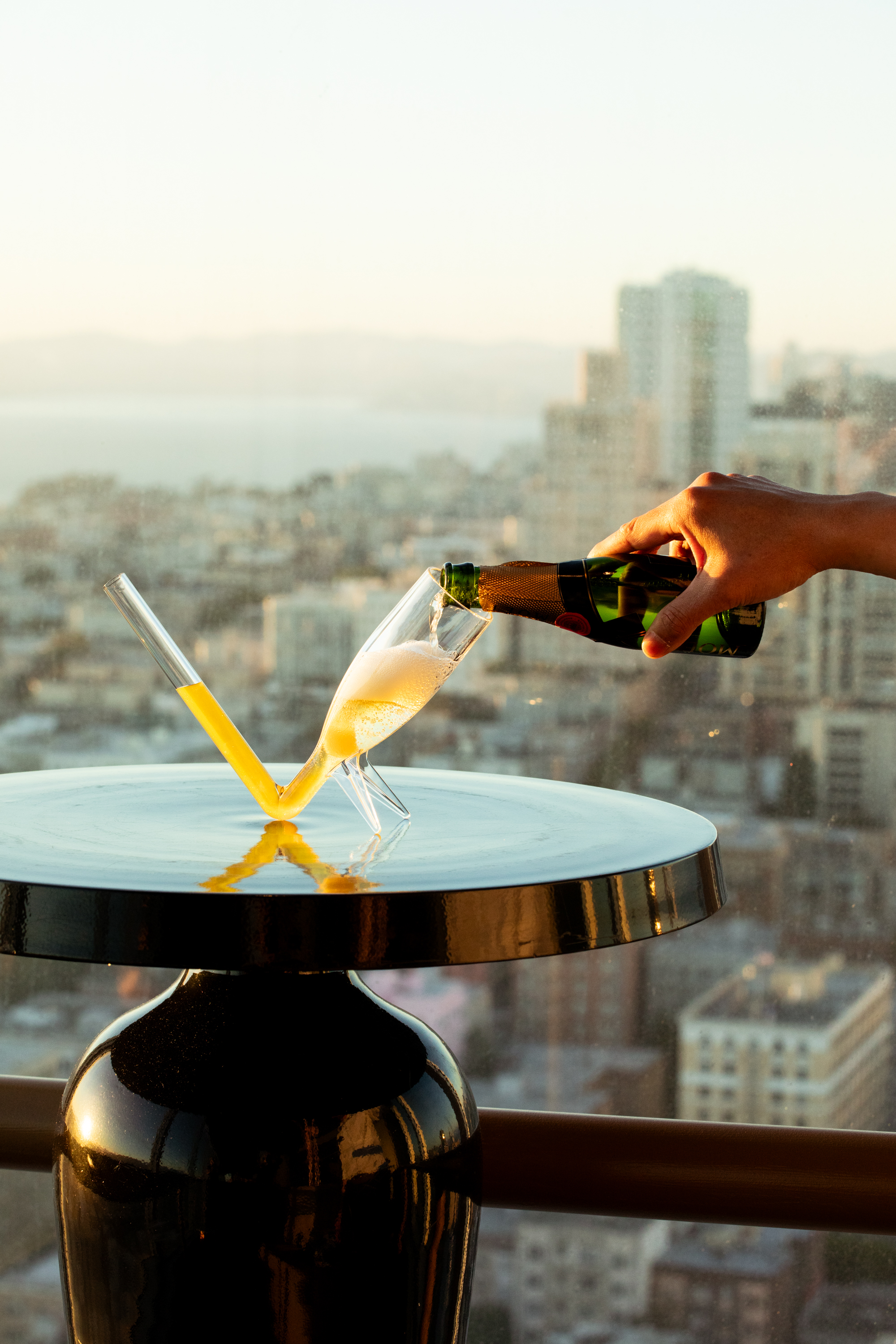A hand pours champagne into a tilted glass on a shiny round table. Next to it, a flute-like glass with yellow liquid stands amid a cityscape backdrop.