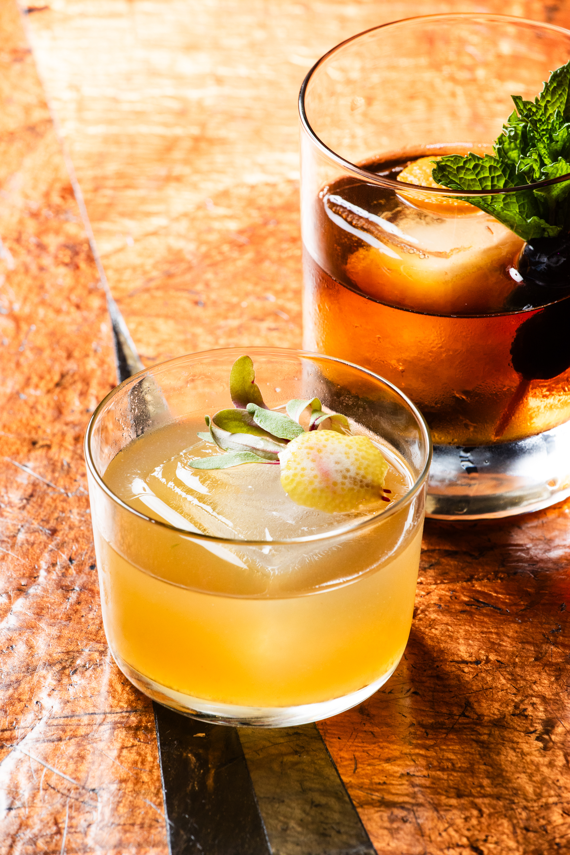 Two cocktails sit on a wooden table: one light-colored with a lemon and herb garnish, and another darker drink with ice, mint, and a garnish.