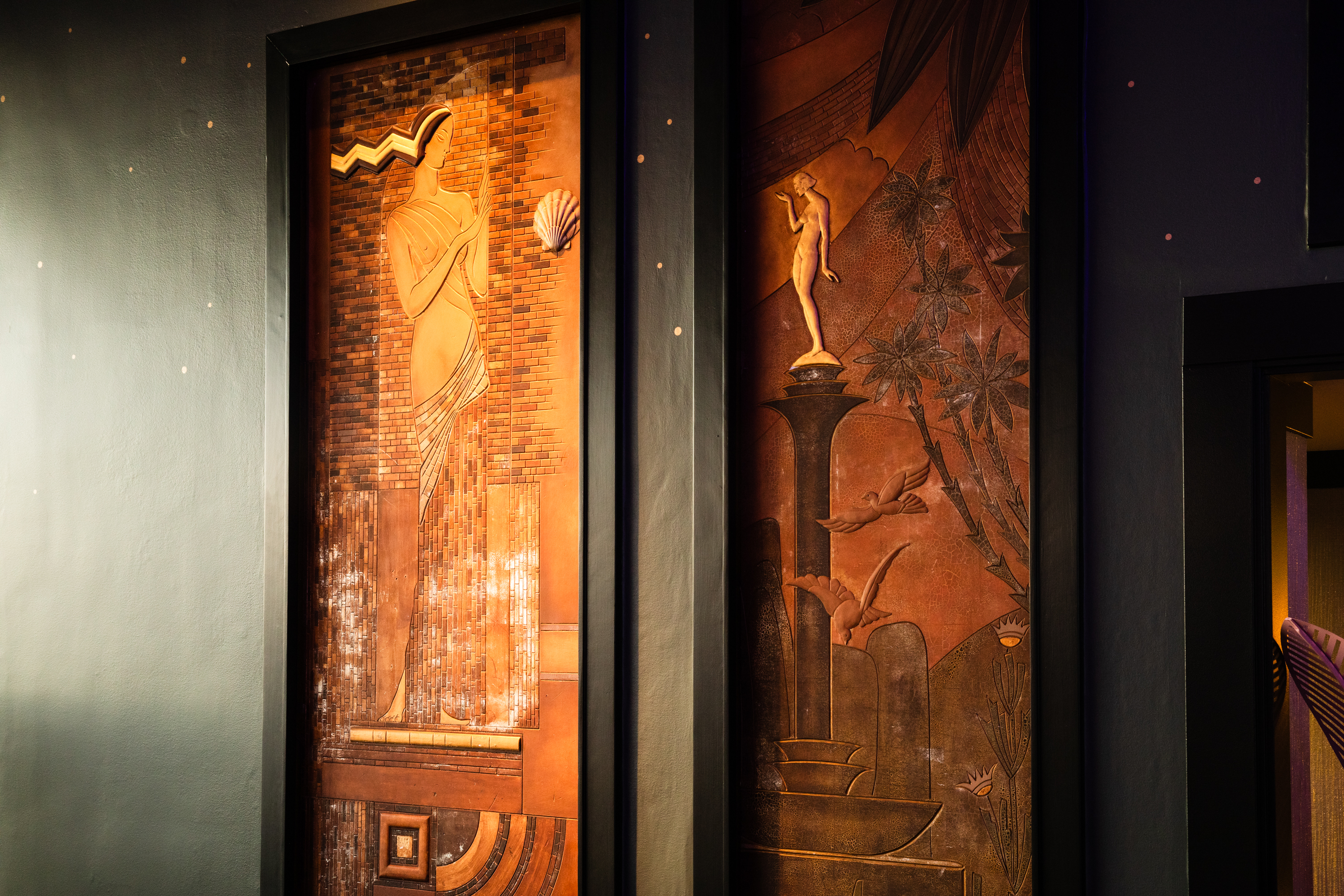 The image shows two vertical, artistic wall panels with intricate carvings. The left panel depicts a stylized figure and patterns, while the right features nature elements and nude figures.