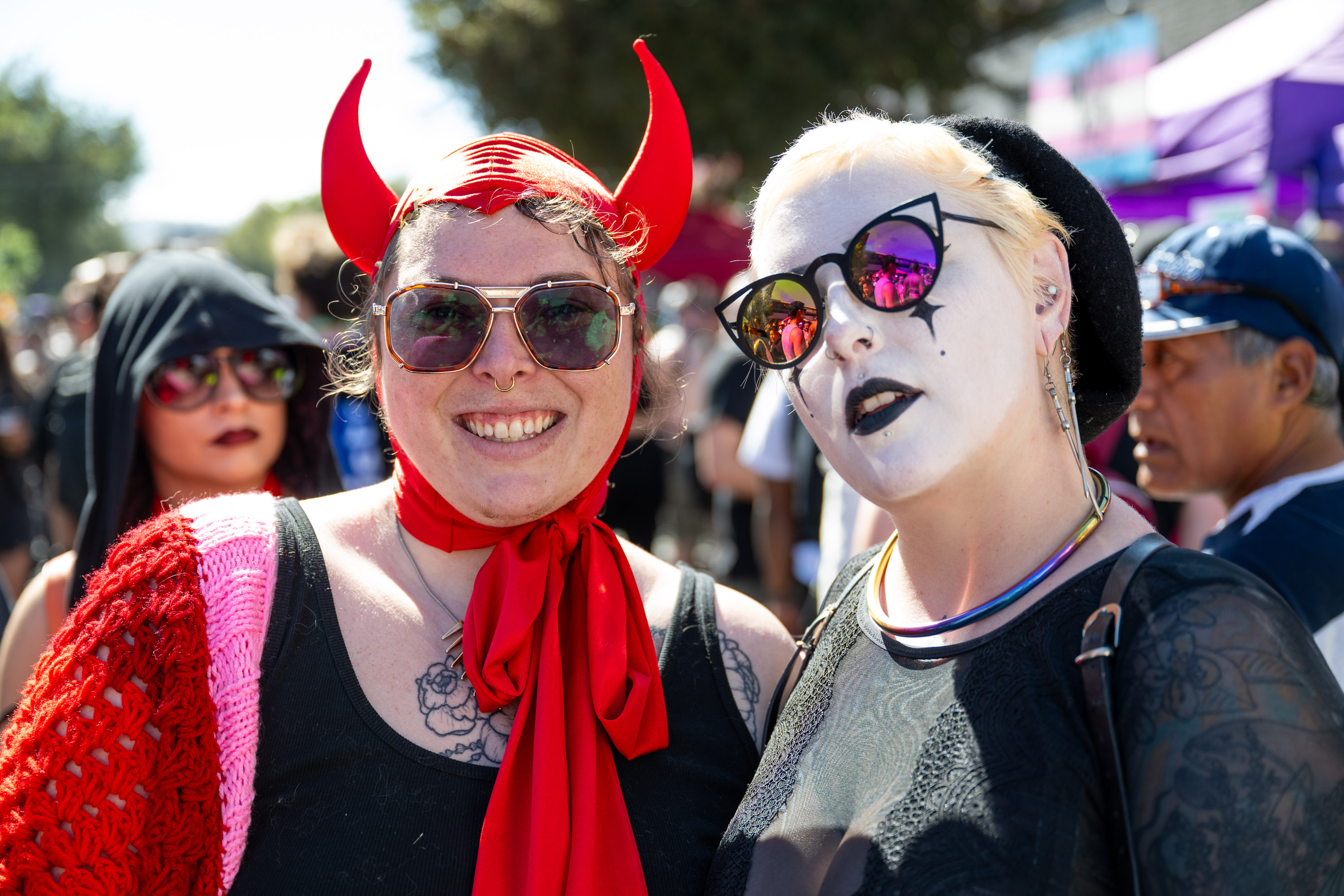 Two people are posing together, one in a devil horn headpiece and glasses, the other in gothic makeup and stylish sunglasses, with a crowd in the background.