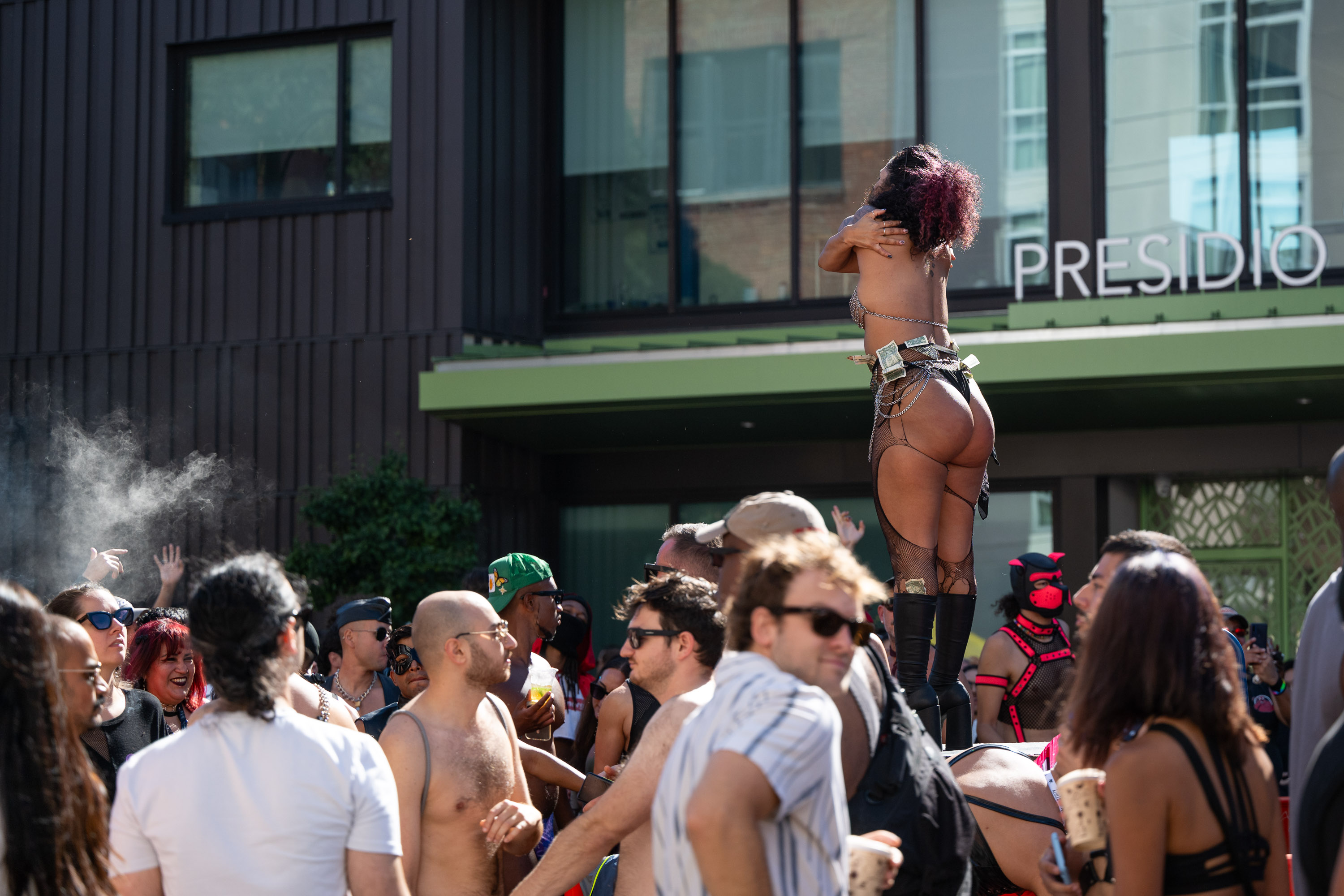 A crowd of people gathers around a performer in revealing attire, standing elevated with money tucked into her outfit, against a backdrop of a modern building with the sign &quot;PRESIDIO.&quot;