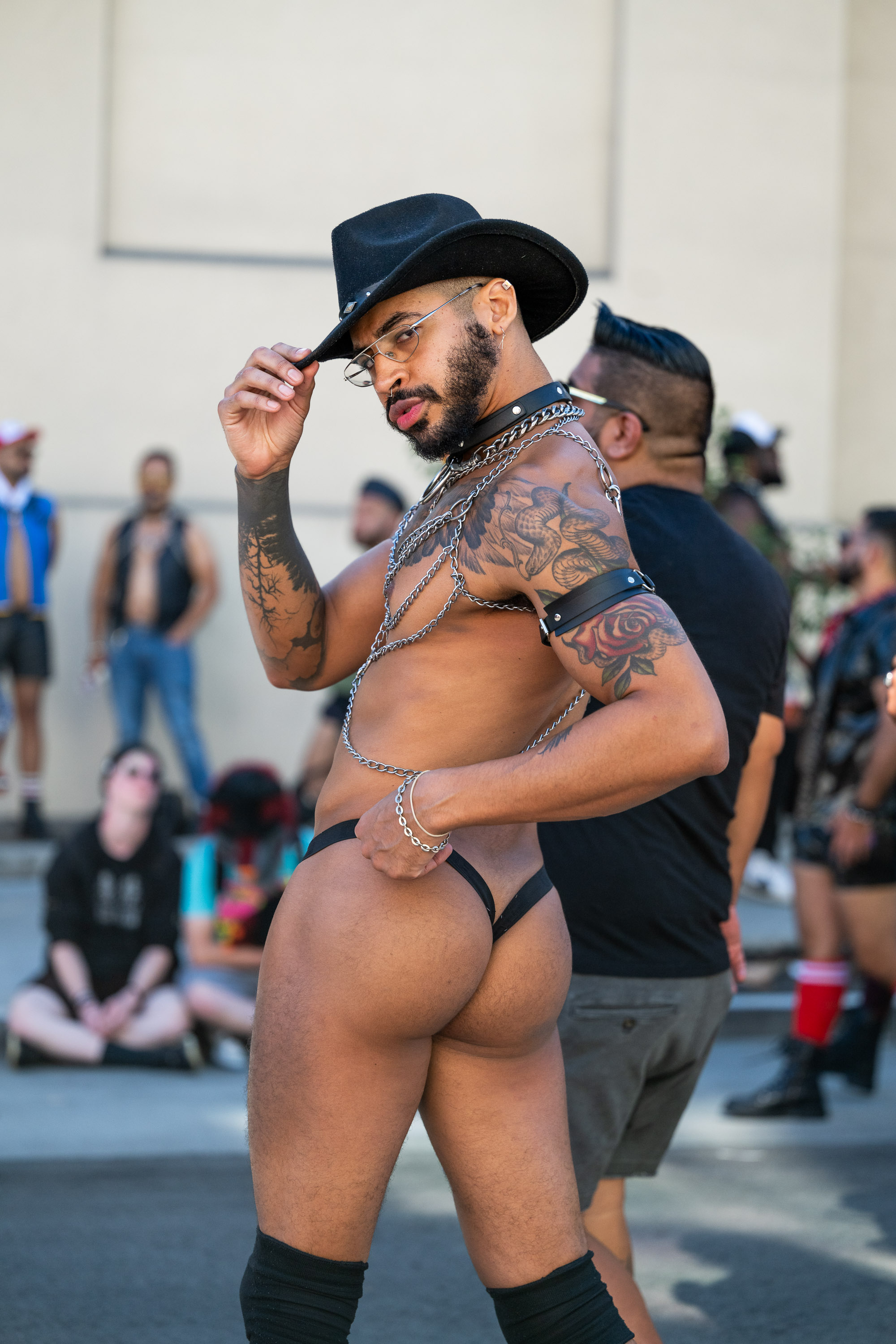 A bearded man in a thong, cowboy hat, and chains poses confidently, looking towards the camera, surrounded by people in casual, eclectic outfits.