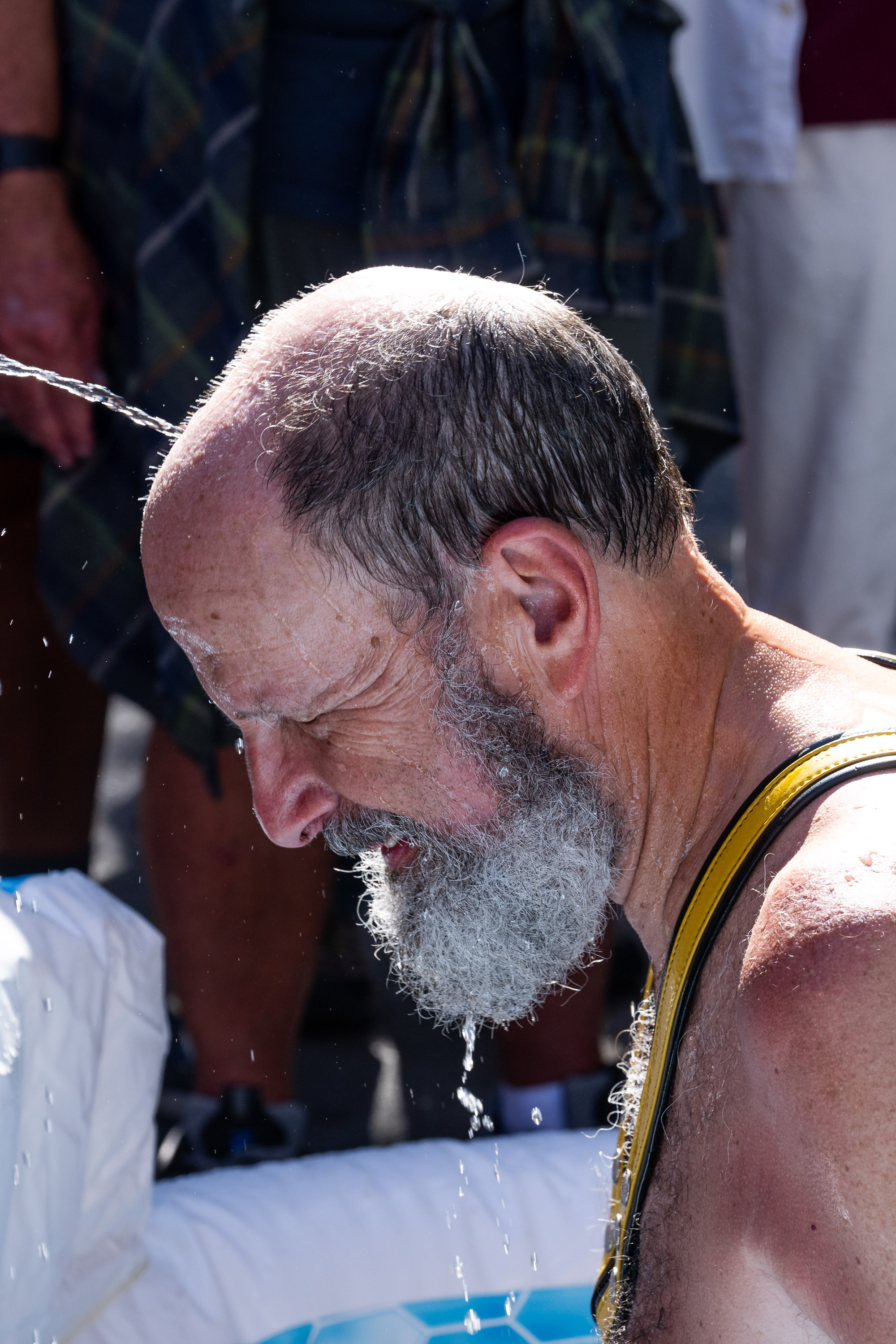 A bearded man with a bald spot is getting his head doused with water, which drips down his face. He is wearing a yellow and black tank top and appears to be outdoors.