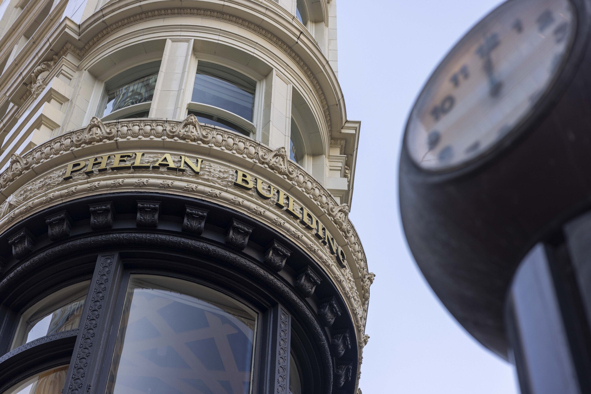 The image shows the ornate upper corner of the Phelan Building, featuring arched windows and intricate architectural details, next to a blurred clock face.