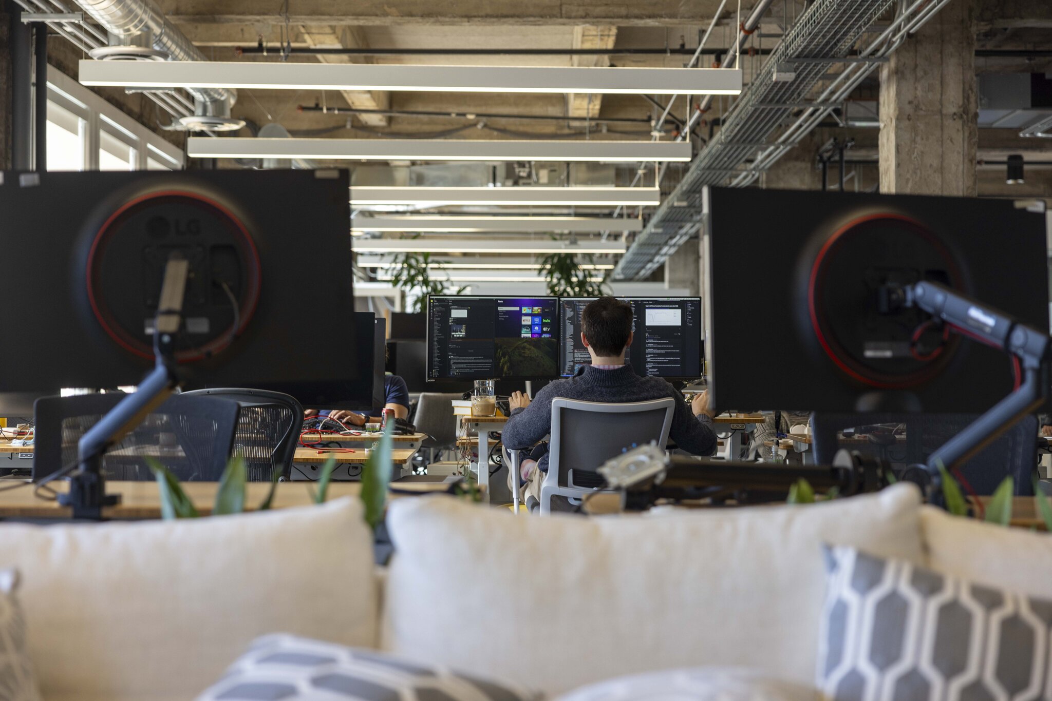 A person is working at a desk in an open-office setting, facing dual computer monitors with code and interfaces displayed, surrounded by modern decor and industrial elements.