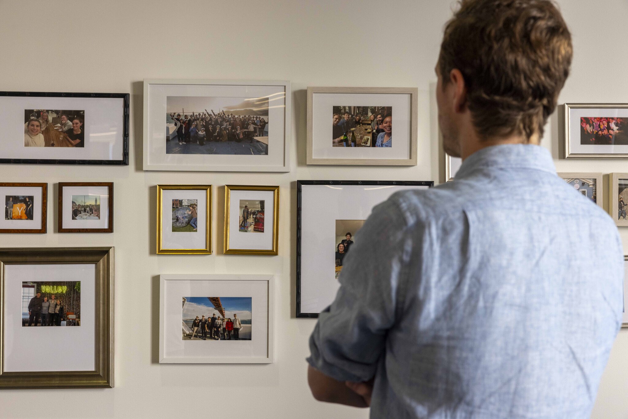 A person in a light blue shirt is looking at a wall with various framed photographs. The pictures depict groups of people and seem to capture different moments and events.