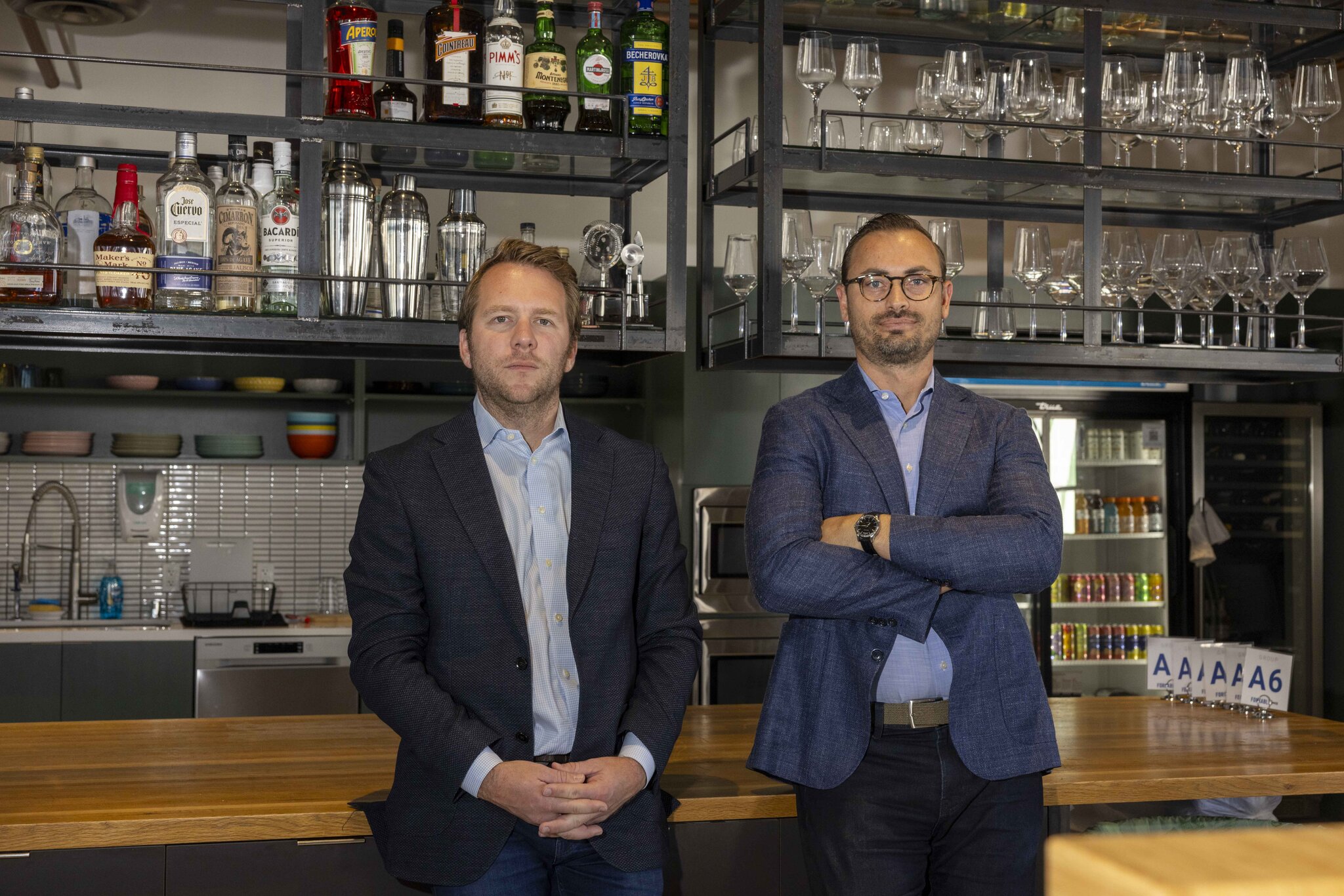 Two men in suits stand in a bar with various liquor bottles, glassware, and a kitchen behind them, posing with serious expressions.