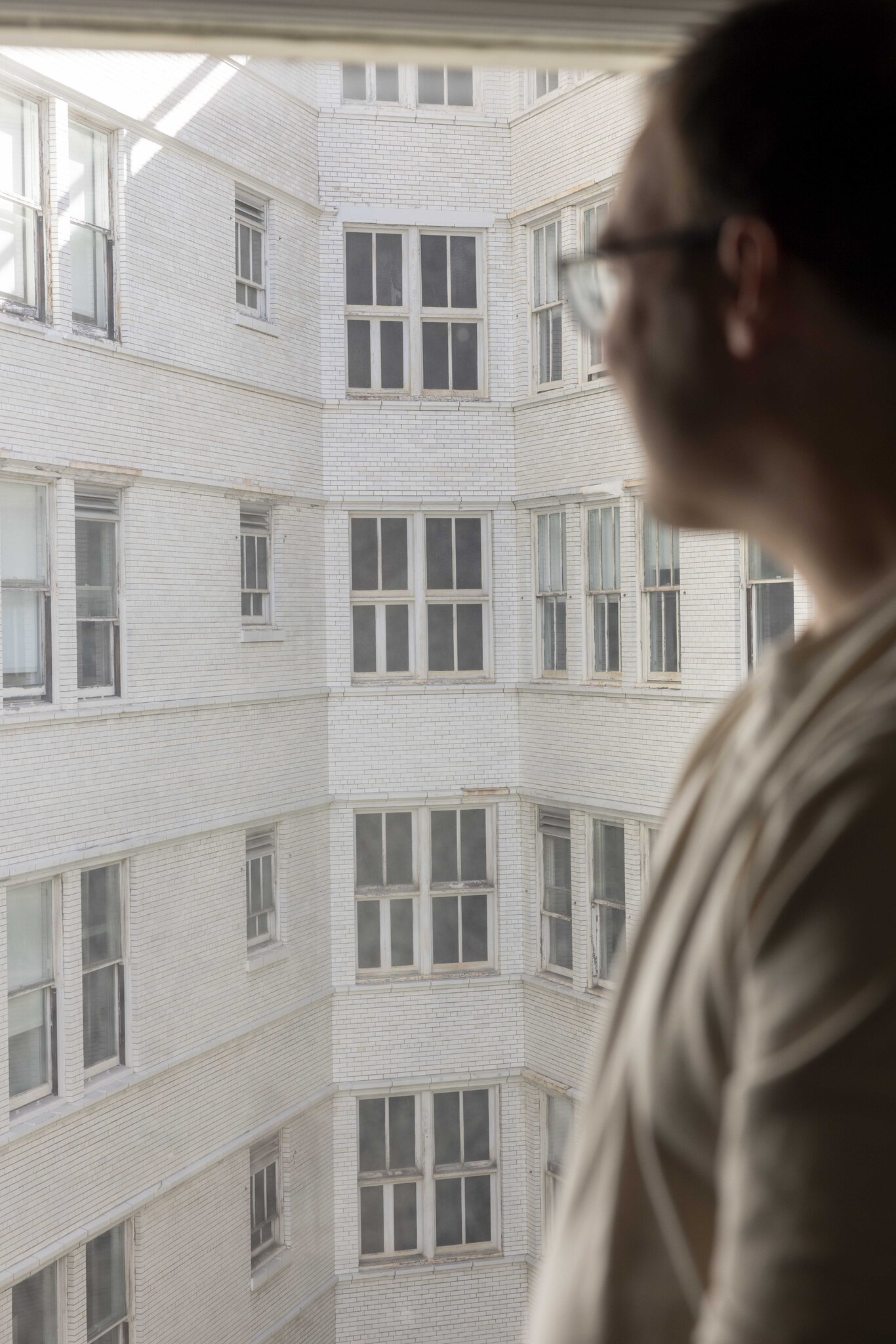 A person in glasses and a beige shirt is seen in profile, looking out of a window at an adjacent white brick building with several rows of closed windows.