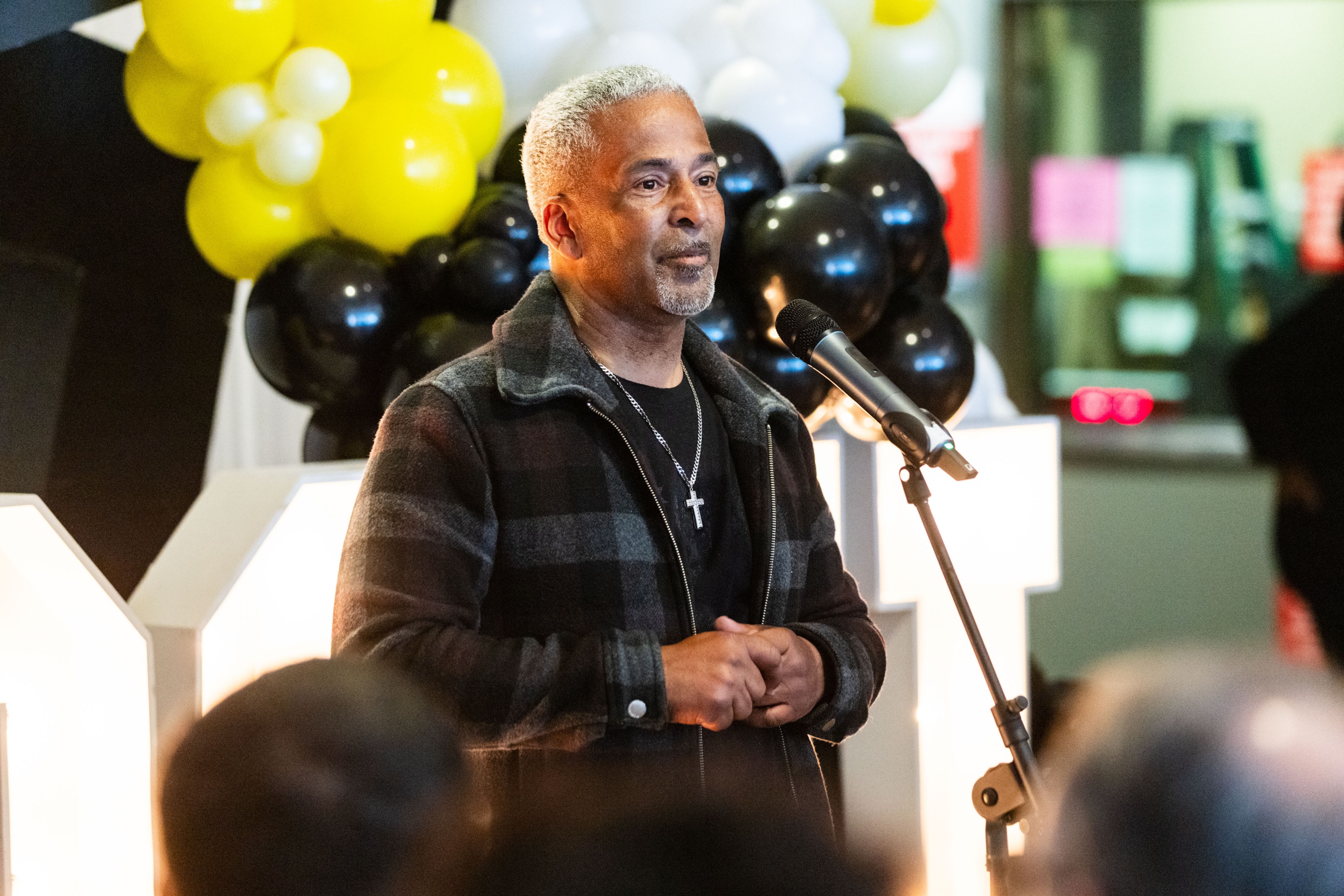 A man speaks into a microphone, standing in front of black, white, and yellow balloons with a calm facial expression, wearing a checkered jacket and a cross necklace.