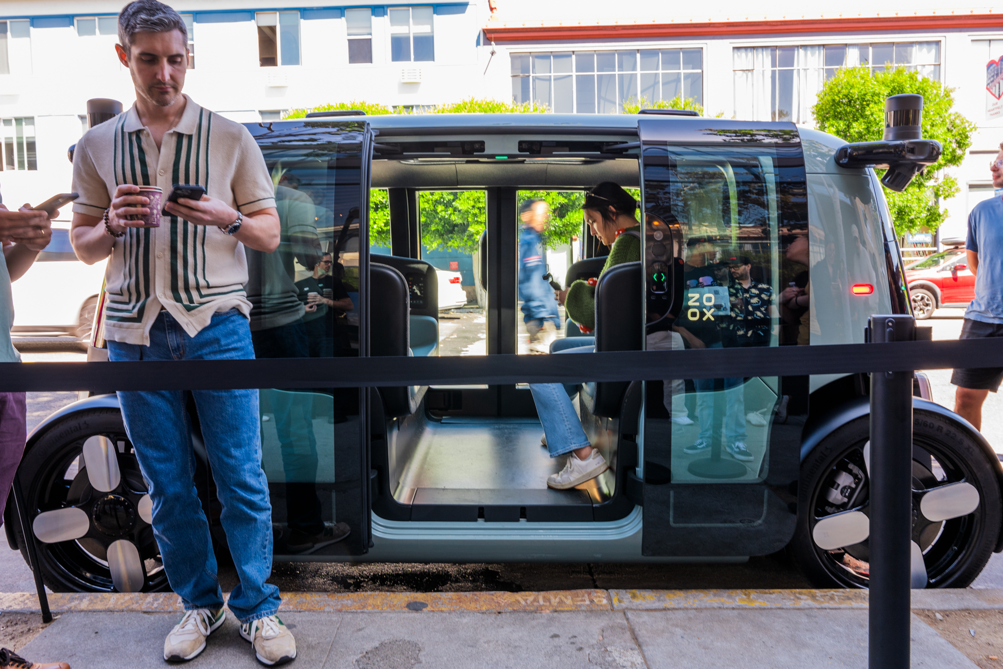 A small group of people are gathered around an autonomous vehicle with sliding doors open, one person inside. A man in front holds a drink and checks his phone.