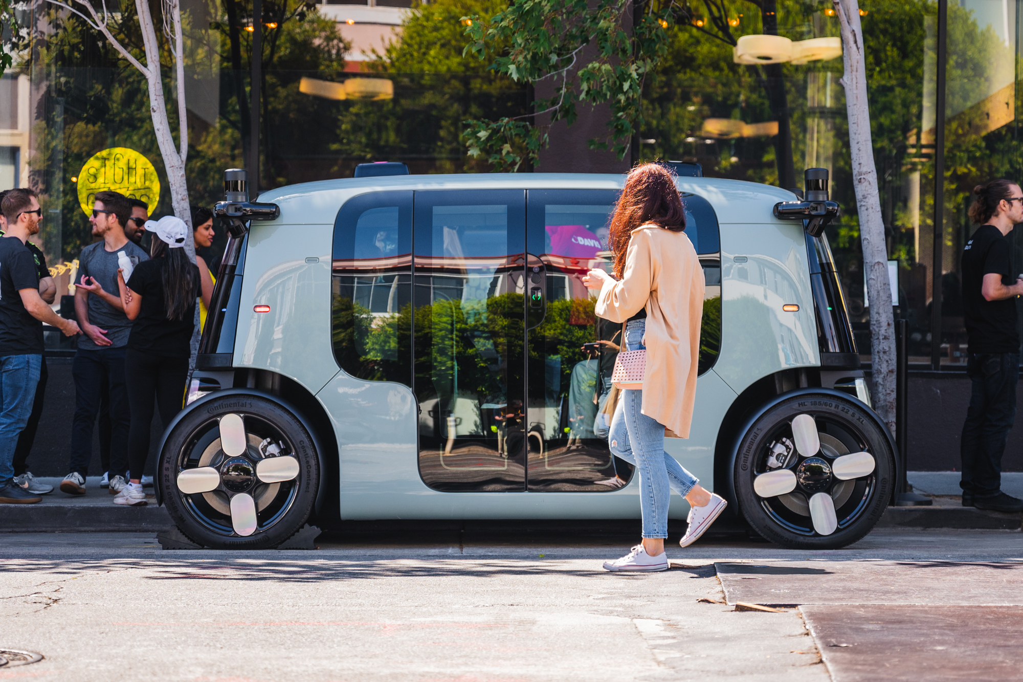 A futuristic, autonomous vehicle is parked on a street. Several people are gathered nearby, and a woman in a long coat and jeans walks by using her phone.