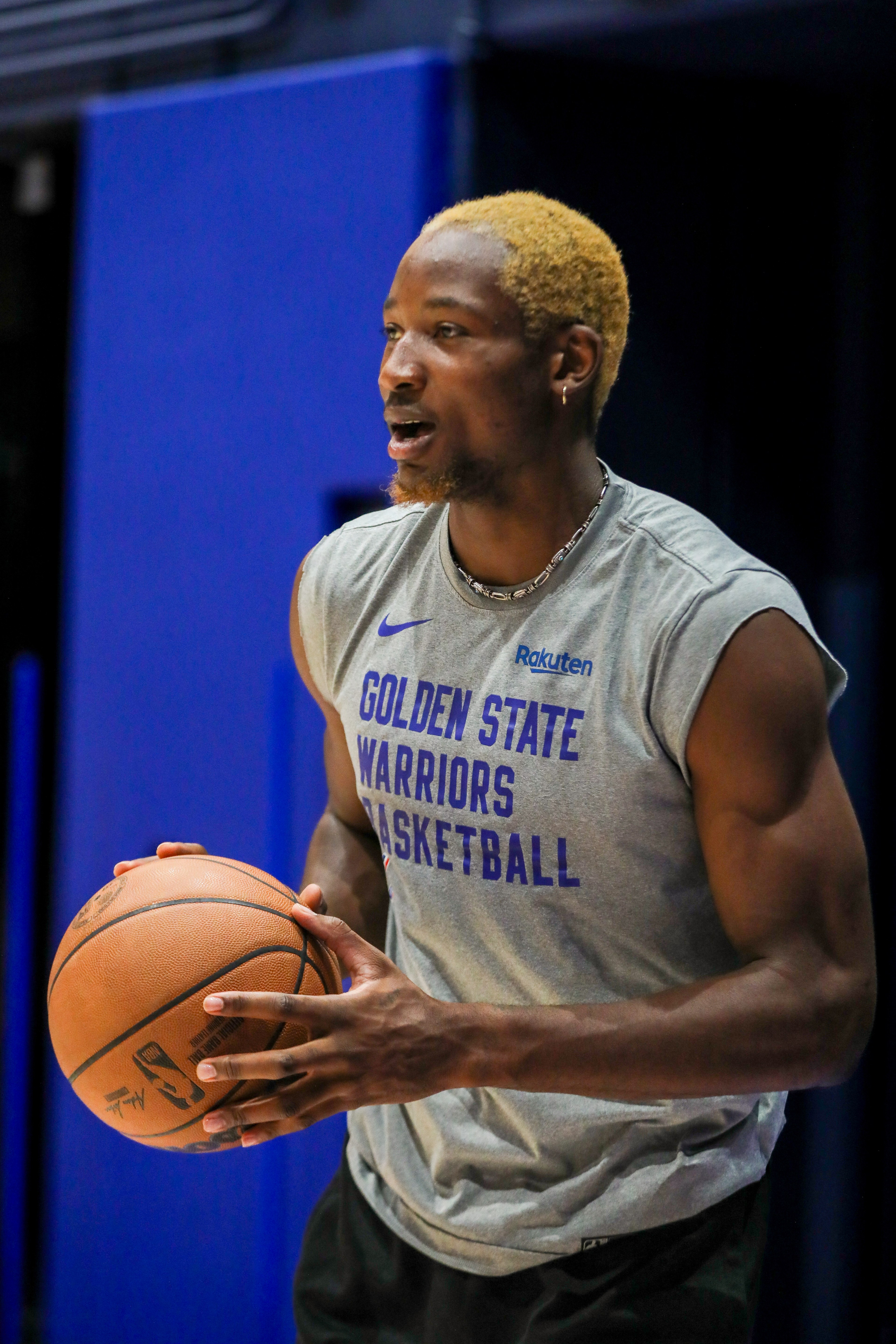 A basketball player with bleached hair holds a ball, wearing a gray "Golden State Warriors Basketball" sleeveless shirt, with a blue background behind him.