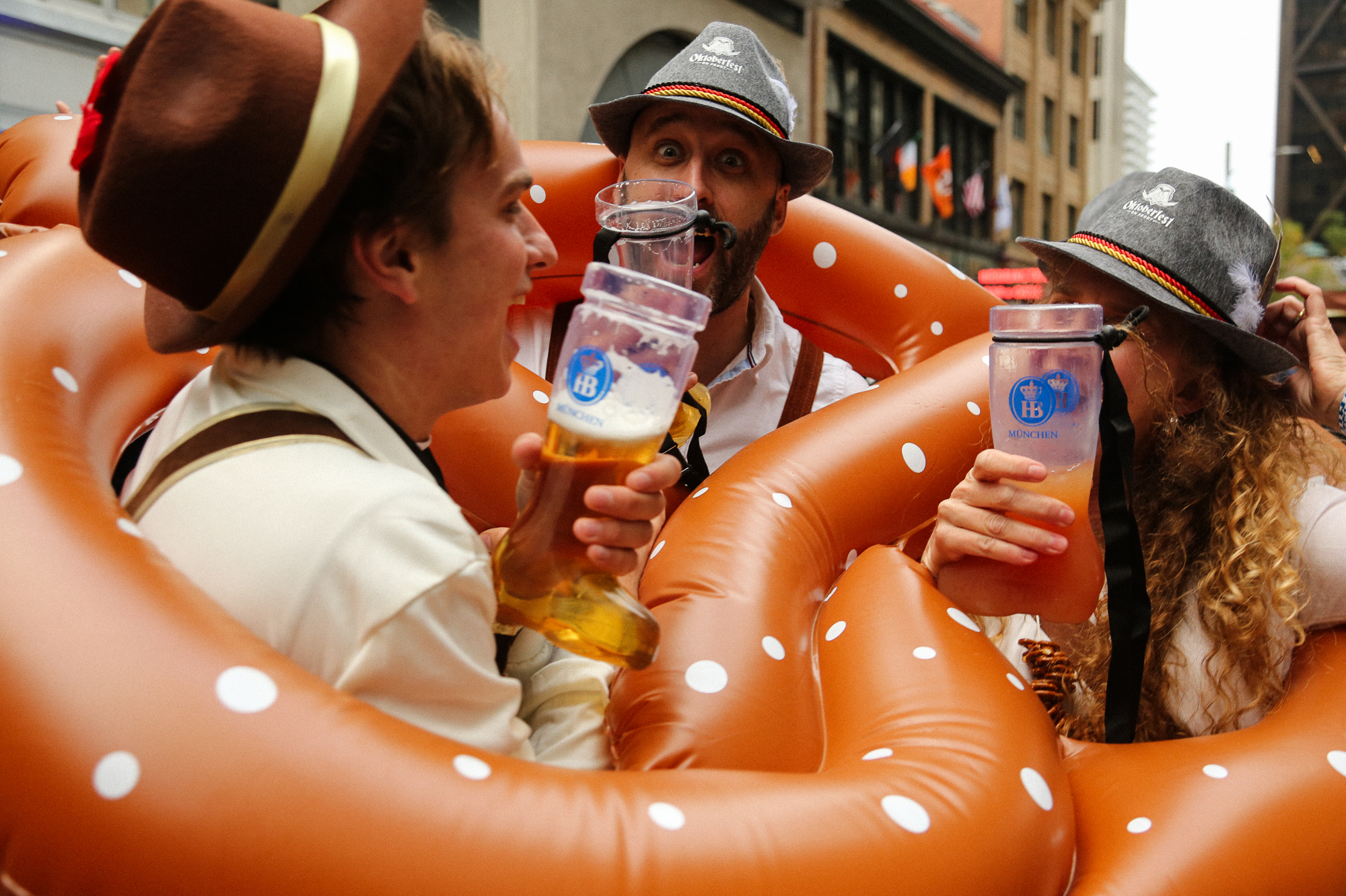 Three people in festive attire drink from large beer glasses, surrounded by inflatable pretzels, celebrating together in an outdoor setting.