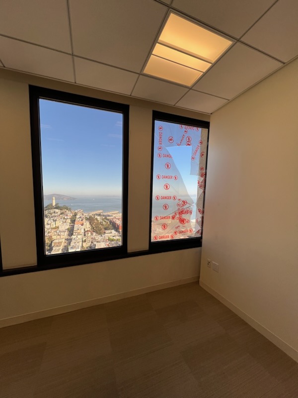 The image shows a room with two large windows; one has a clear view of a cityscape and the ocean, while the other is covered in red "DANGER" stickers.