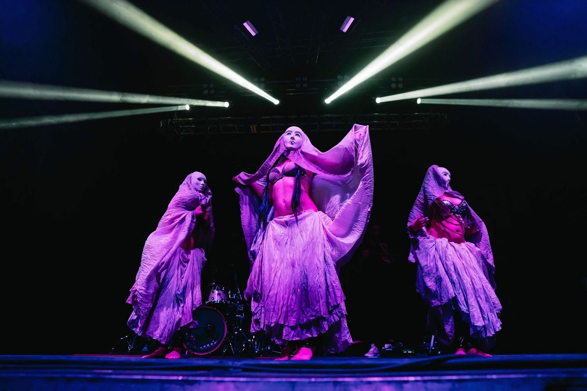 Three dancers on a dimly lit stage wear long, flowing skirts and veils. Spotlights shine from above, illuminating their vibrant costumes and theatrical poses.