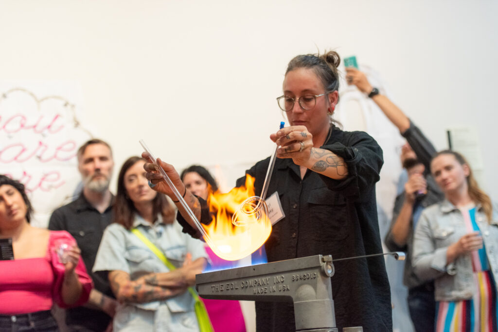 A woman demonstrates glass-blowing, holding rods over a flame, while a group of onlookers closely watches her. The background features a neon sign saying &quot;you are here&quot;.