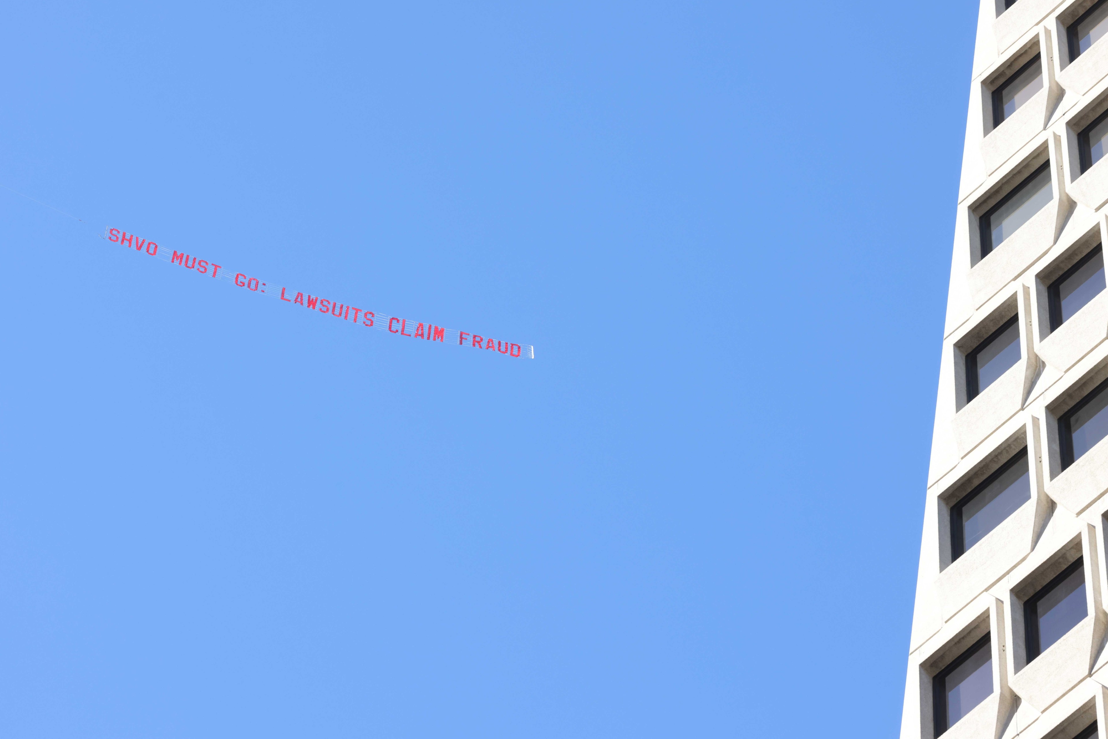 A banner towed by an aircraft reads, &quot;SHVO MUST GO: LAWSUITS CLAIM FRAUD,&quot; against a clear blue sky, with a corner of a modern, windowed building visible on the right.