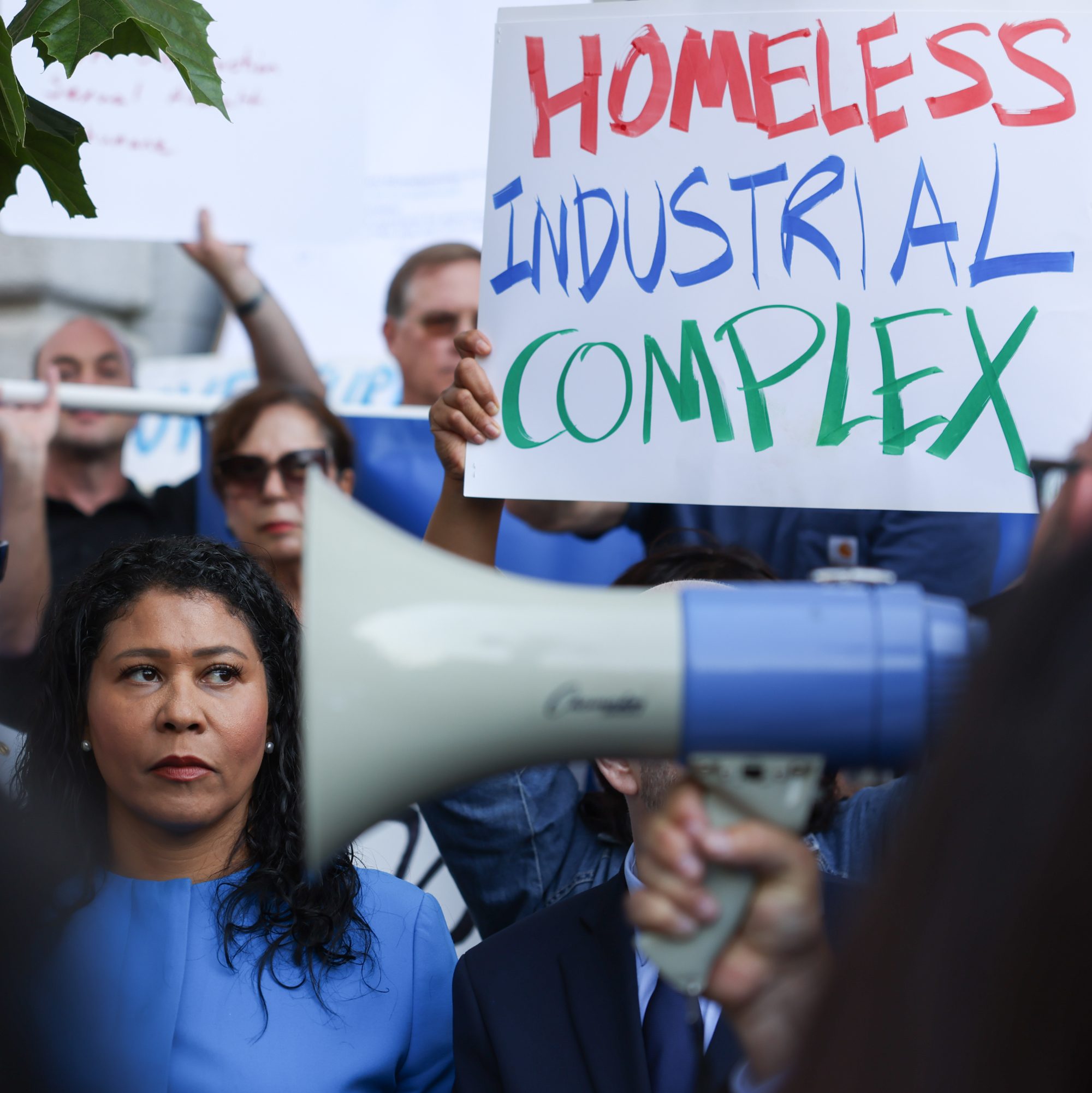 A woman with a megaphone is in focus with protesters and a &quot;HOMELESS INDUSTRIAL COMPLEX&quot; sign in the background.