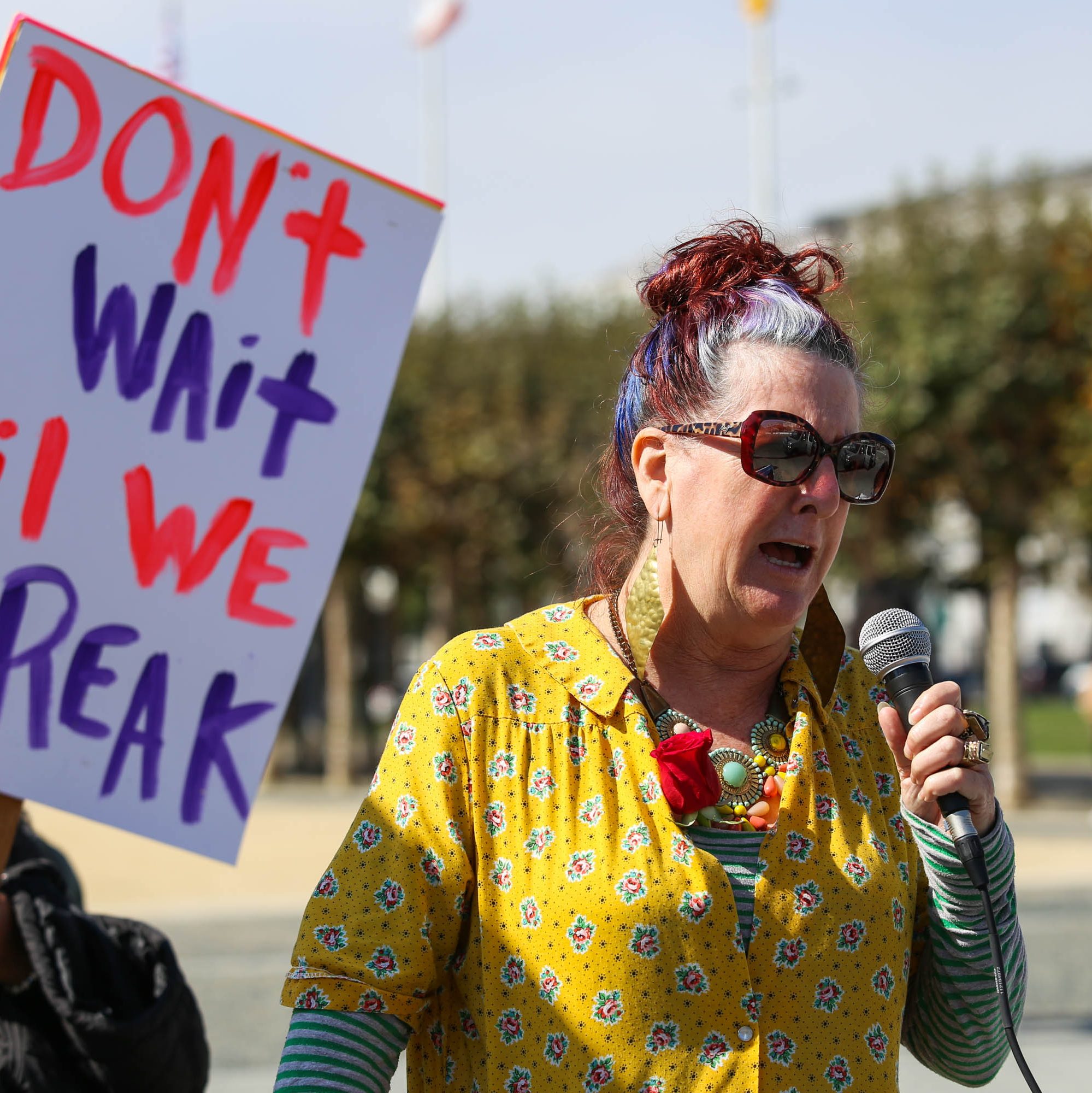 A woman in colorful attire and sunglasses speaks into a microphone. A sign nearby reads, &quot;DON'T WAIT TIL WE BREAK&quot; in bold red and blue letters.