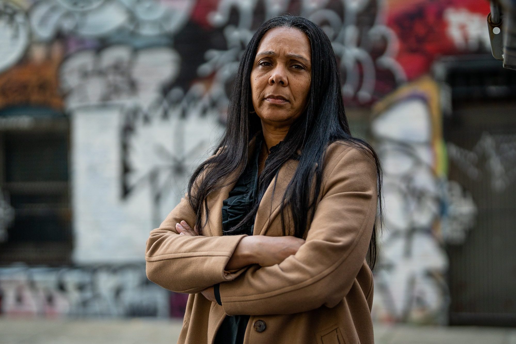 A woman with long dark hair, wearing a tan coat, stands with arms crossed in front of a graffiti-covered wall on an urban street. She has a serious expression.