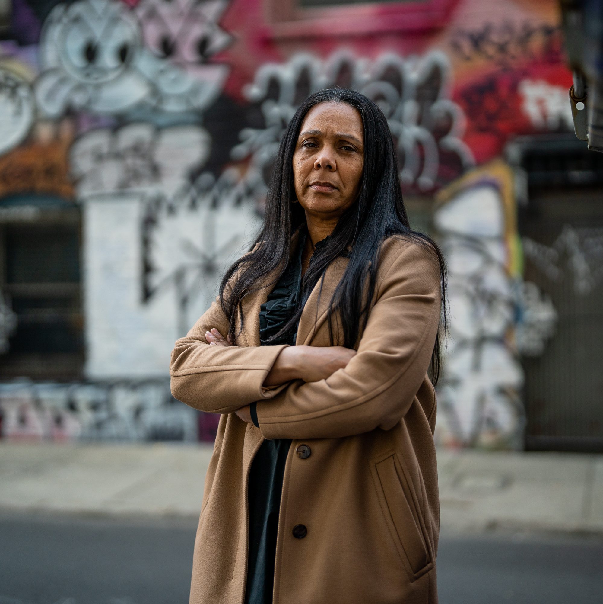 A woman with long dark hair, wearing a tan coat, stands with arms crossed in front of a graffiti-covered wall on an urban street. She has a serious expression.